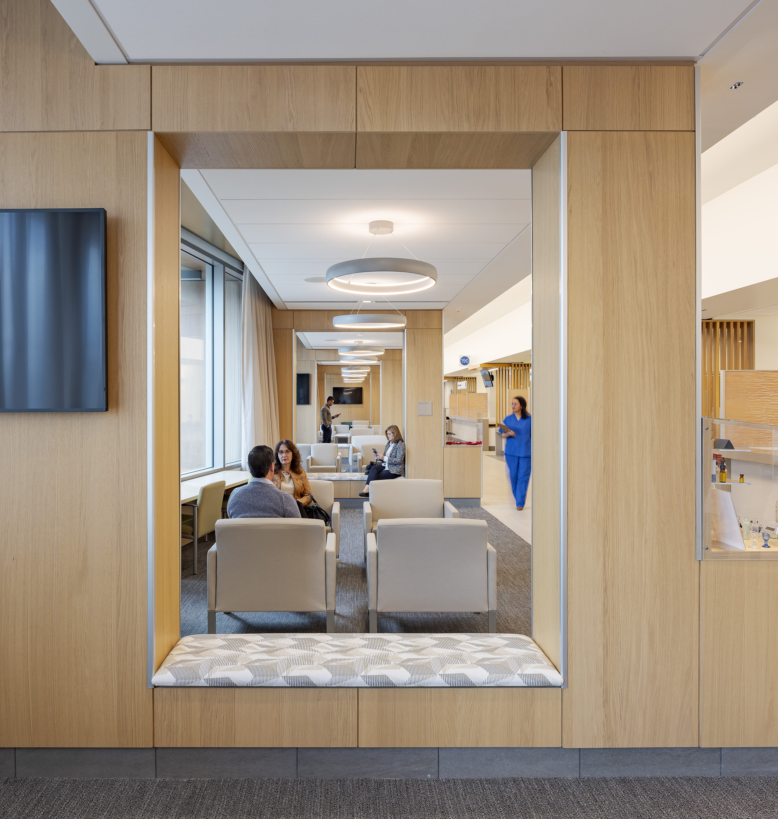 Wood-framed view into a bright waiting area with upholstered seating, circular pendant lights, patients, and a staff member moving through the adjacent clinical corridor.