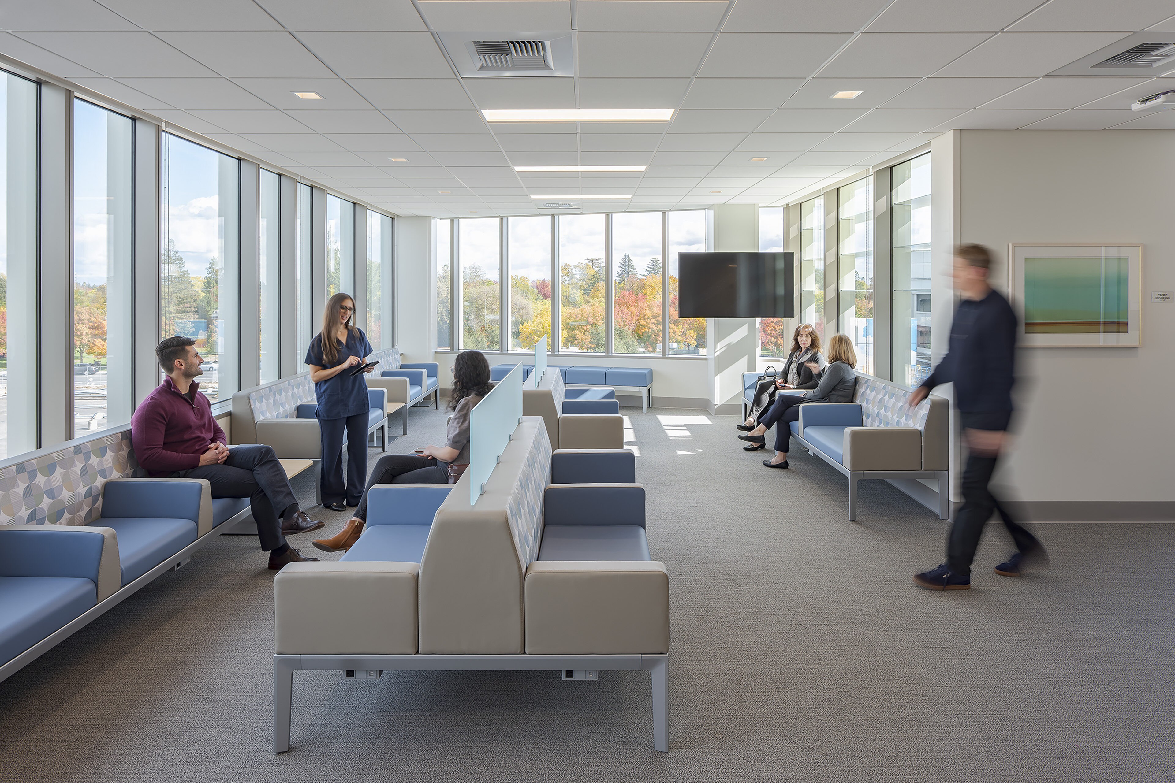 Bright waiting area at the UC Davis Health Eye Institute with floor-to-ceiling windows, blue modular seating, patients, and a staff member assisting a visitor.