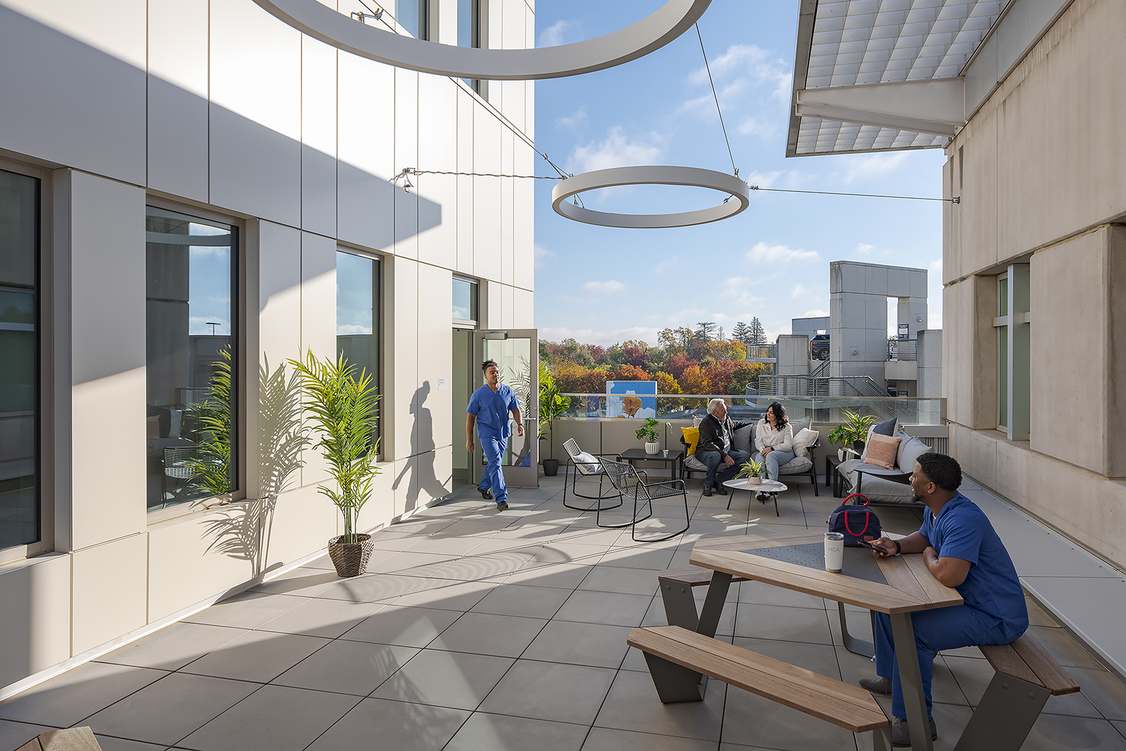 Outdoor terrace with circular suspended sculptures, lounge and picnic seating, potted plants, and patients and staff using the space.