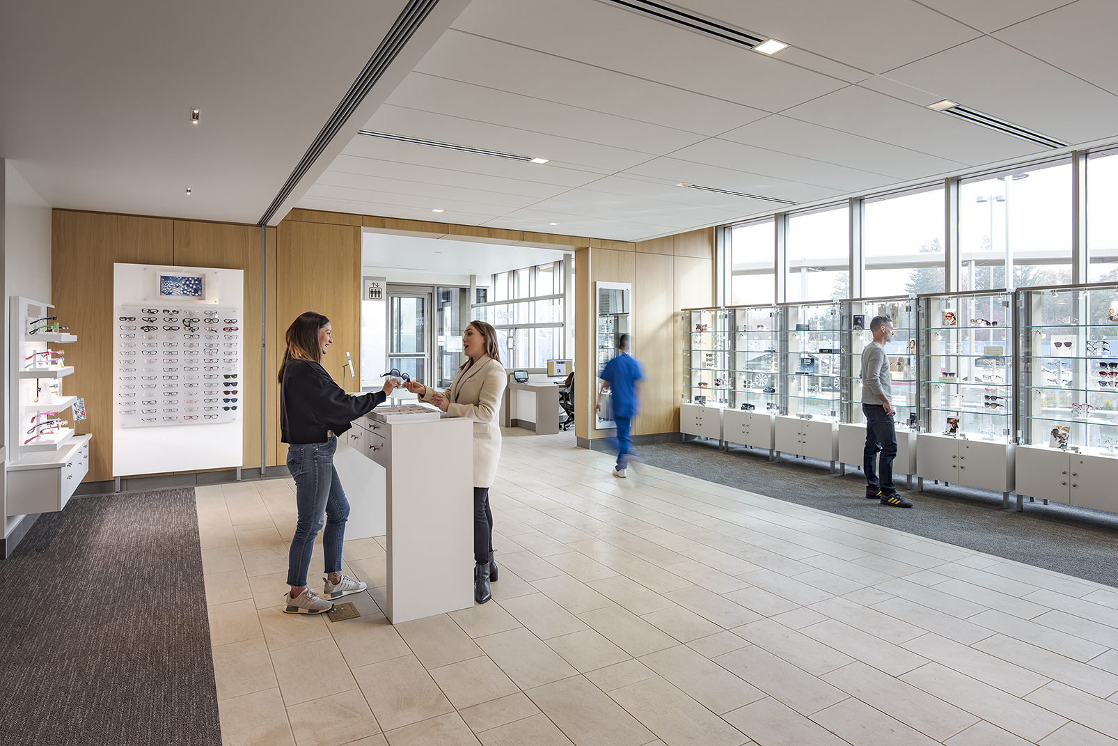 Bright optical retail area with eyewear displays, floor-to-ceiling windows, a central consultation counter, and visitors browsing frames.