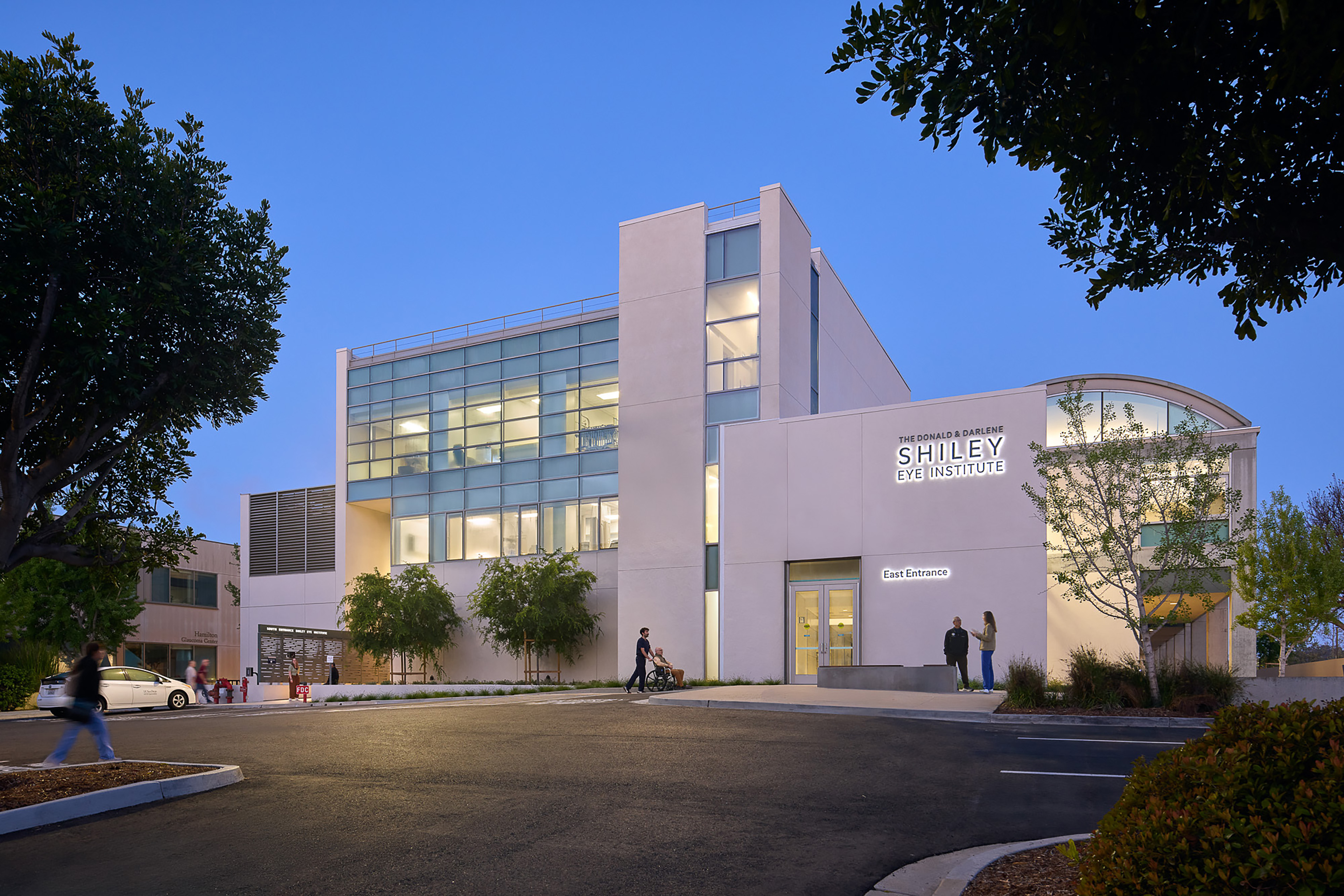 Dusk exterior of the Donald P. and Darlene V. Shiley Eye Institute, showing the illuminated east entrance, glass façade, curved roofline, landscaping, and visitors arriving.