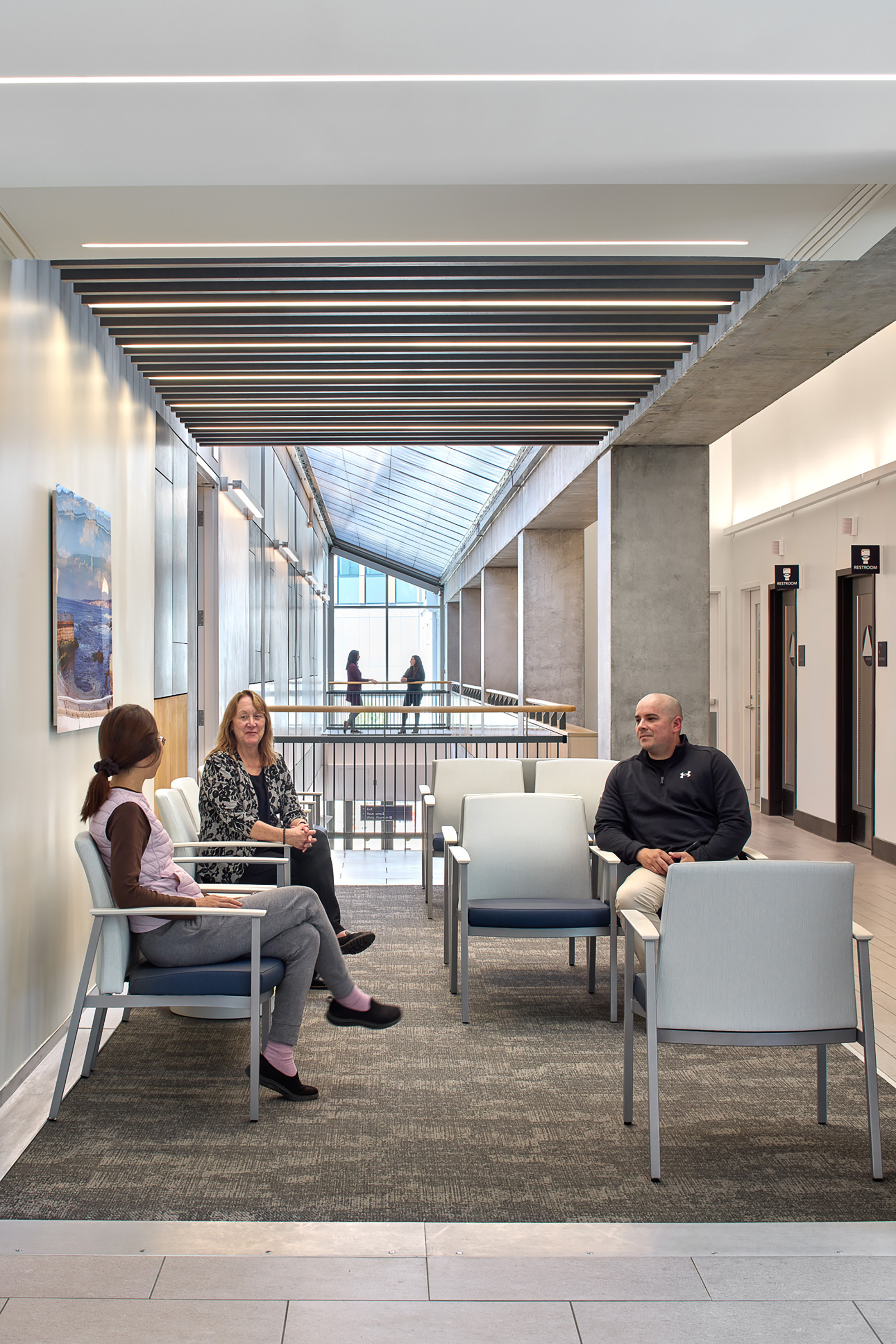 Upper-level waiting area at the Shiley Eye Institute with rows of seating beneath linear wood ceiling slats, exposed concrete columns, a skylit atrium, and visitors seated and conversing.