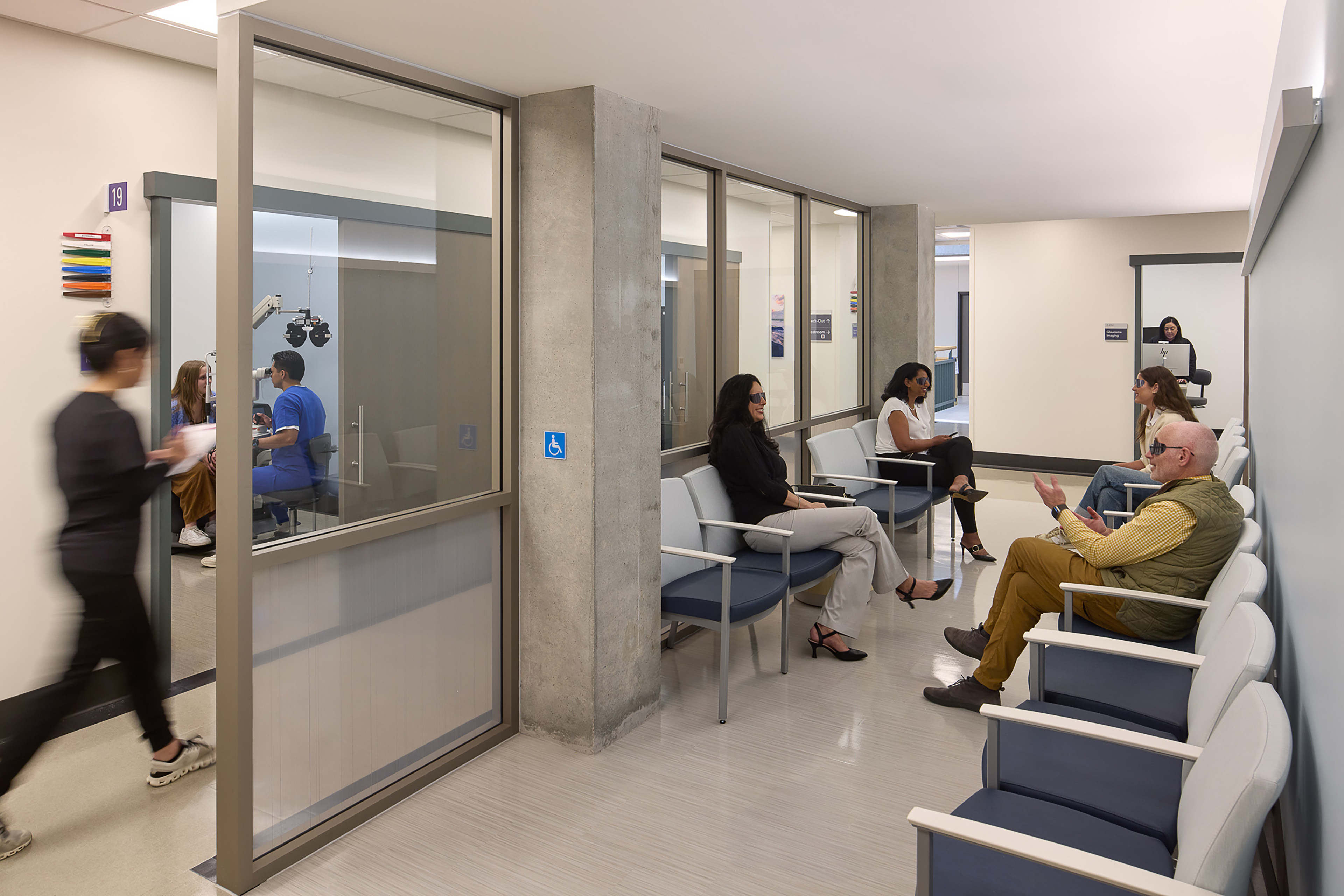 Clinical waiting area with rows of blue-and-gray seating, glass-fronted eye exam rooms, exposed concrete columns, and patients and staff using the space.