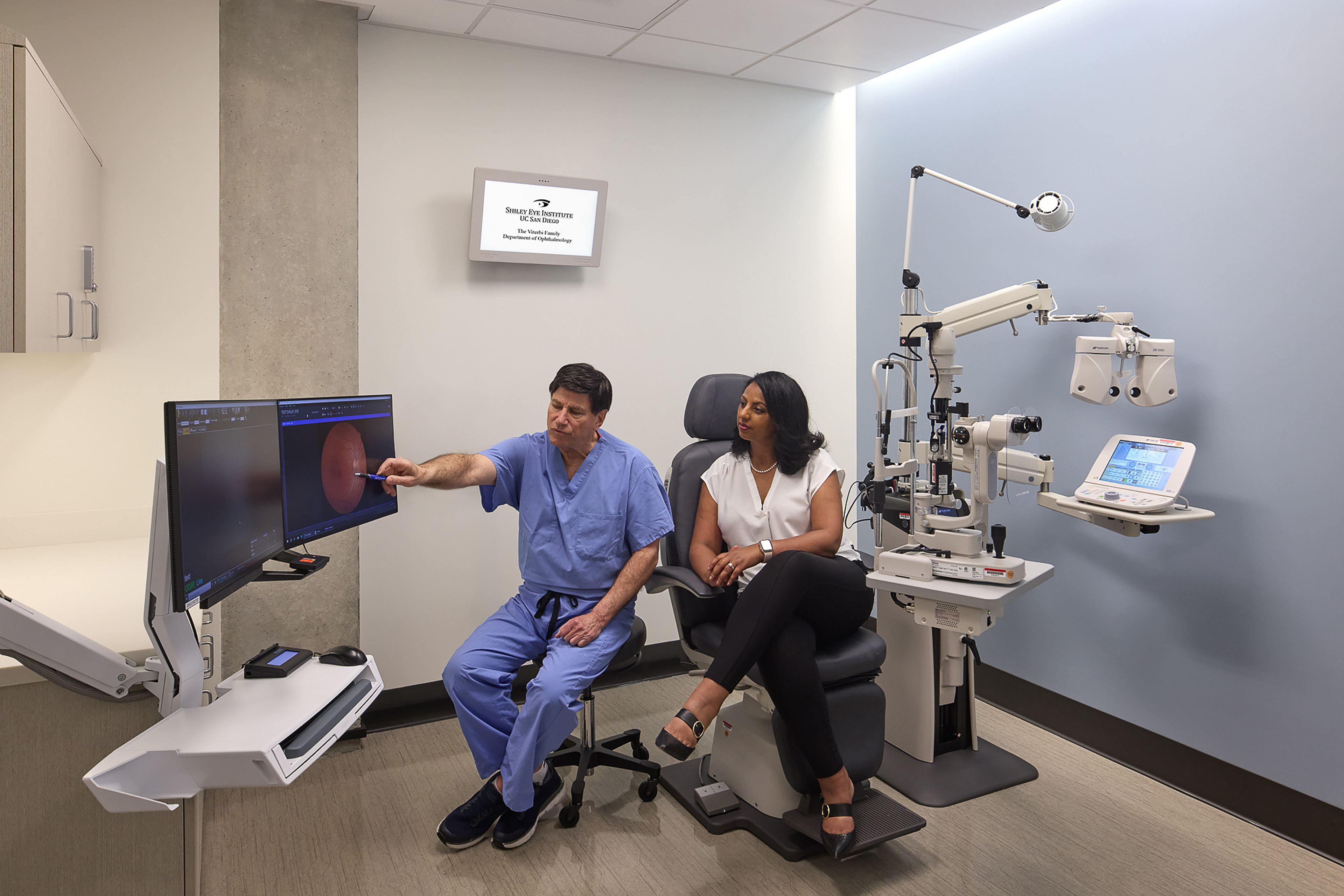 Ophthalmologist reviewing and pointing to a retinal image on a monitor while discussing the results with a seated patient in the exam room.