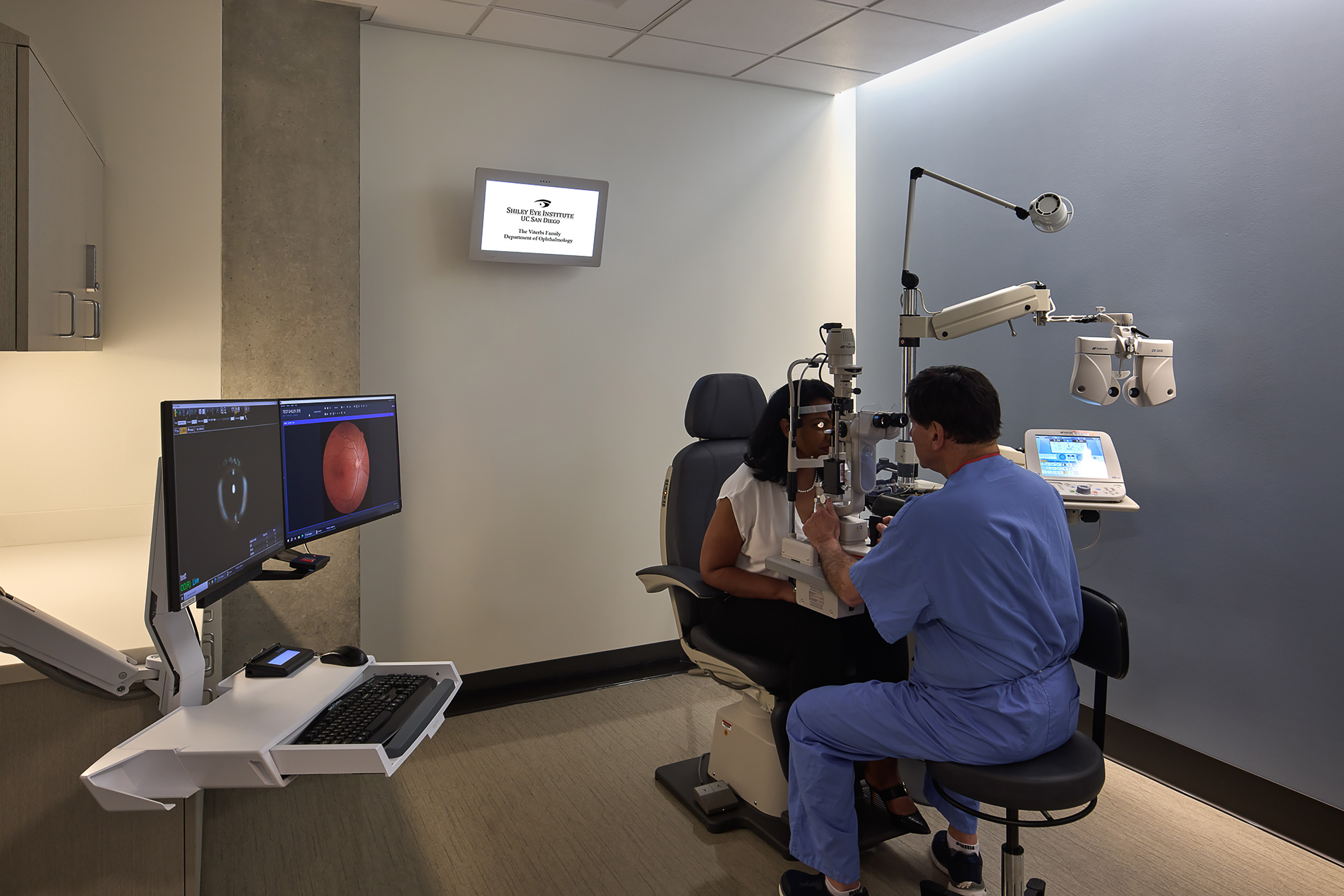 Ophthalmologist using a slit-lamp microscope to examine a patient’s eyes in a modern exam room equipped with imaging monitors and specialized diagnostic equipment.