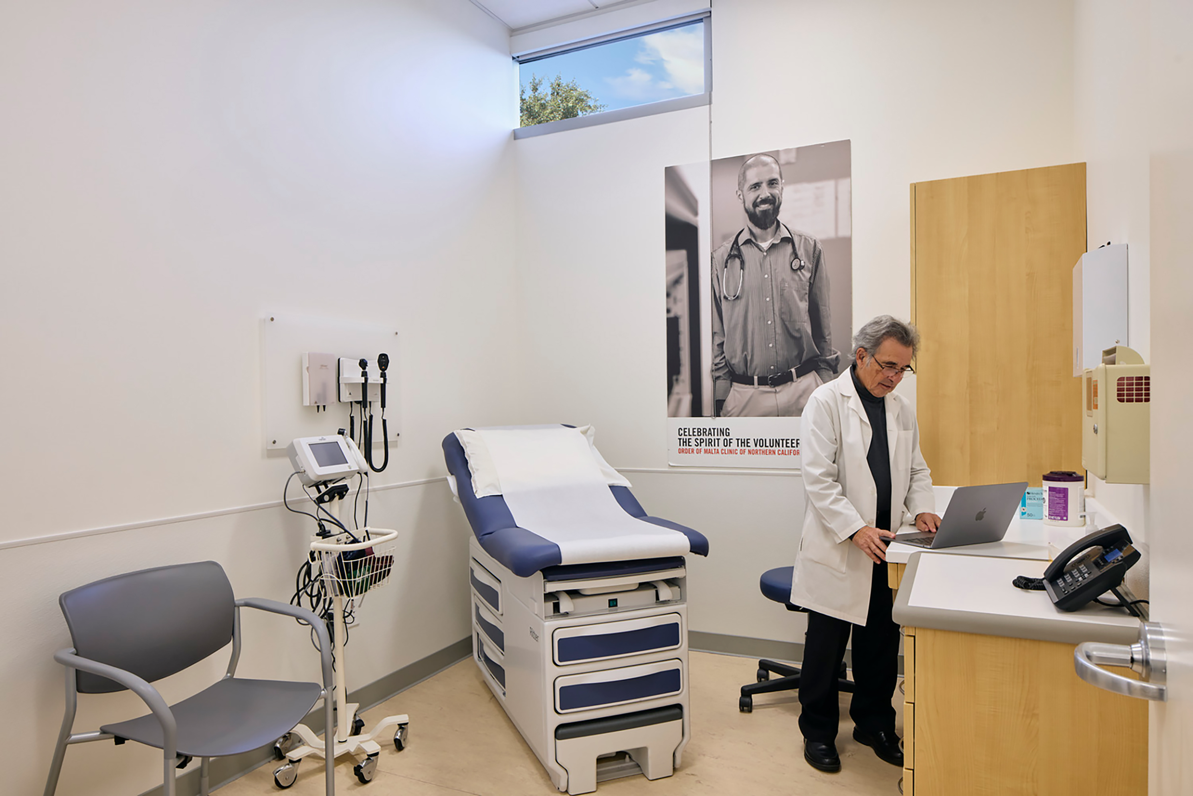 Medical exam room with an examination table and diagnostic equipment, where a physician in a white coat works at a laptop beneath a large portrait of a volunteer doctor.