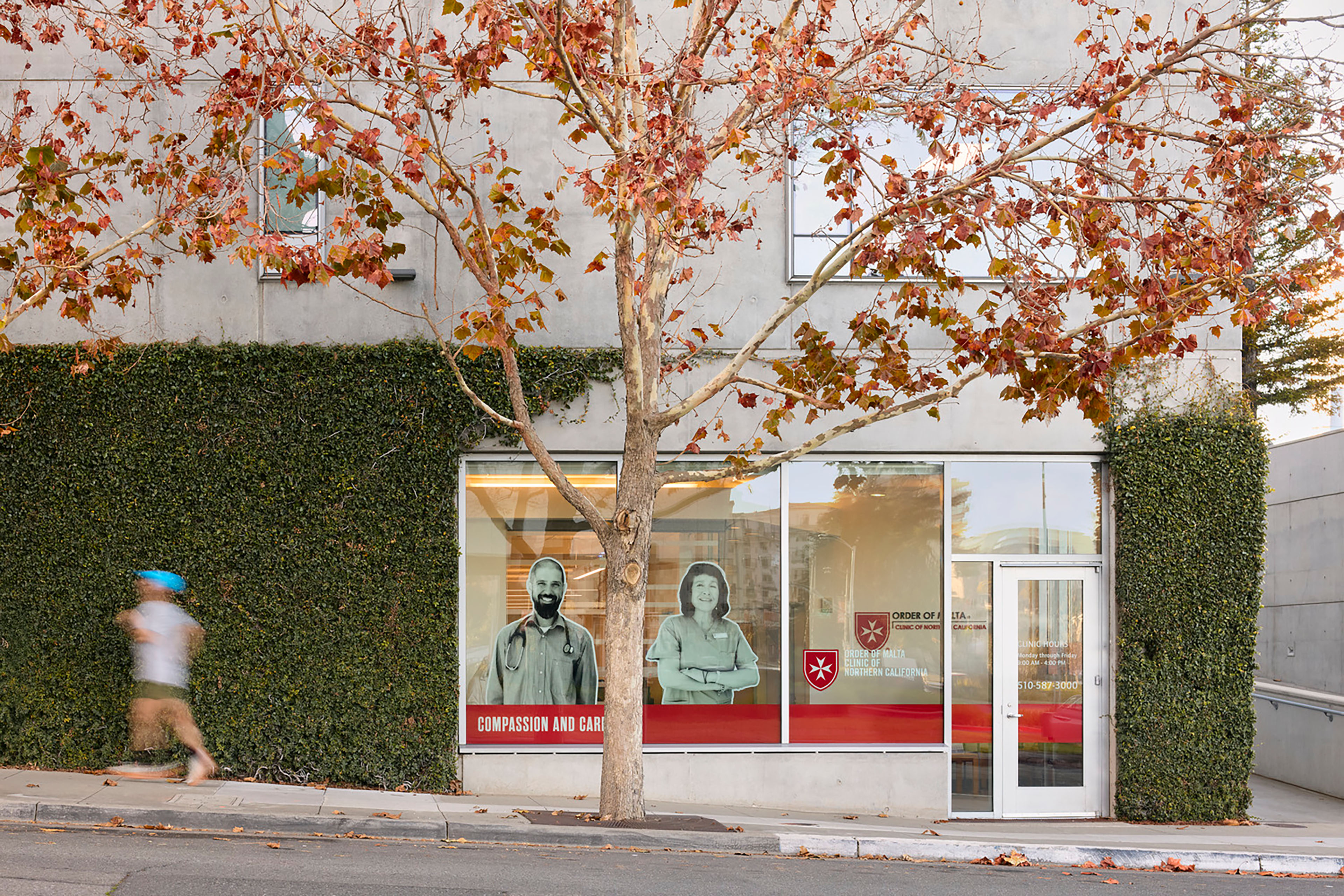 Clinic exterior with a living green wall, autumn tree, storefront graphics featuring healthcare staff, and a pedestrian passing along the sidewalk.