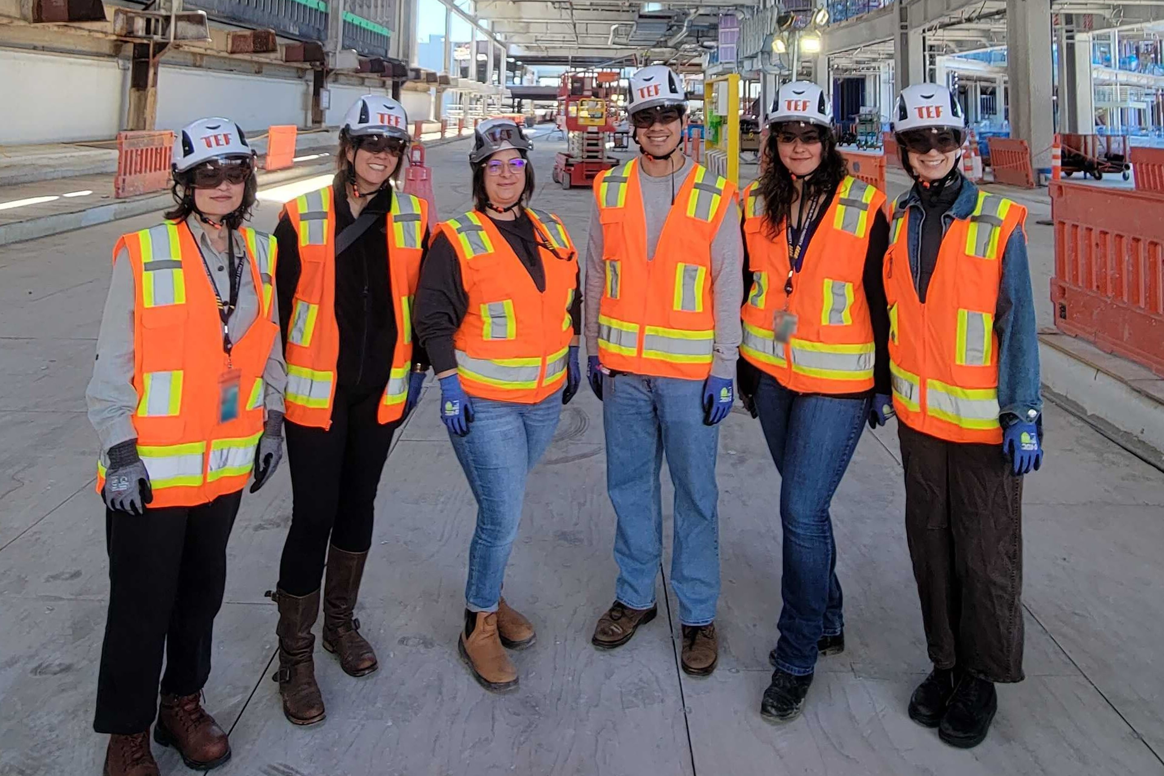 Six TEF team members pose together at an active construction site wearing branded hard hats, safety glasses, gloves, and bright orange reflective vests.