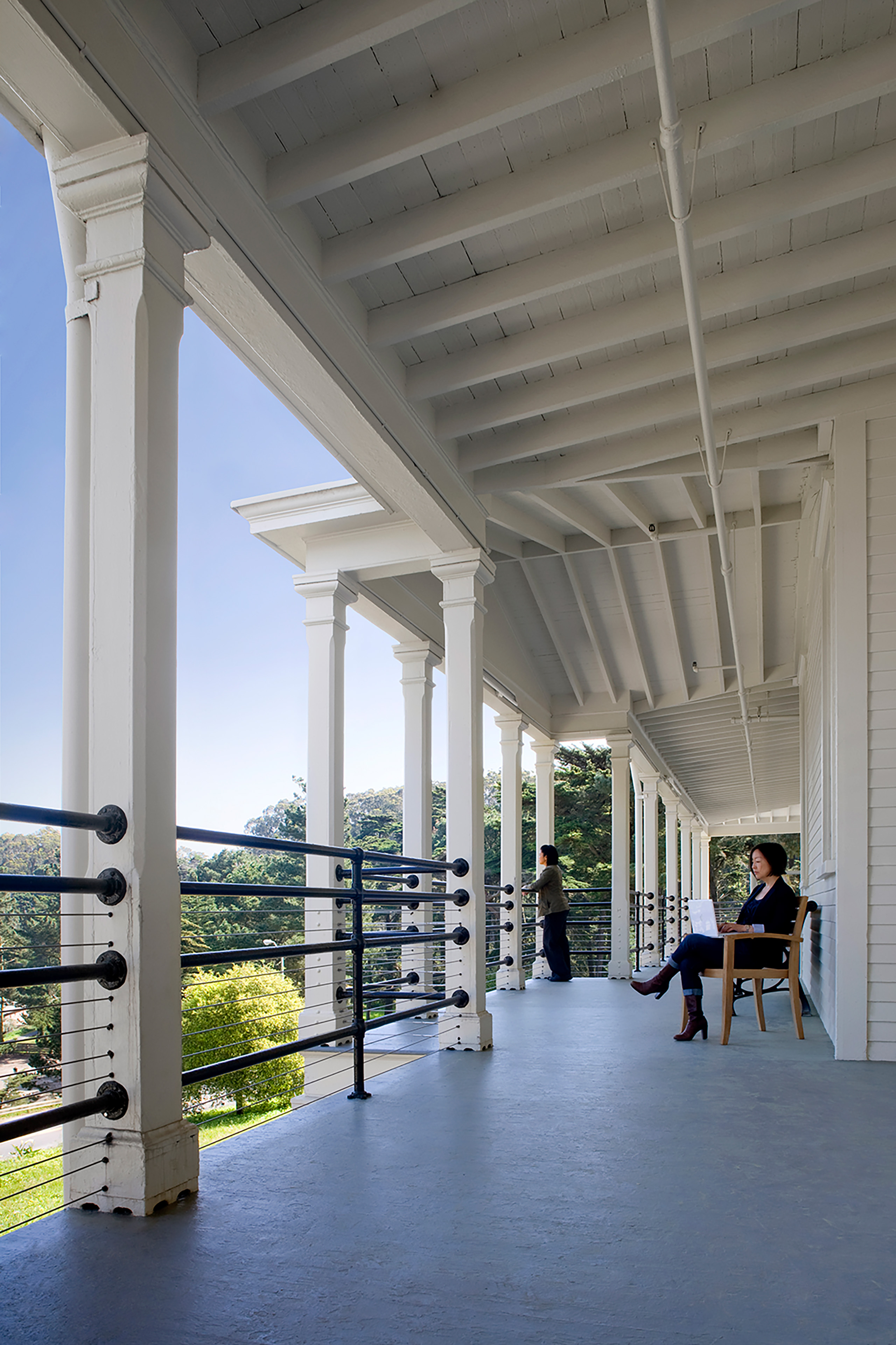 Covered upper-level veranda of a historic white building, with exposed beams, square columns, black railings, one person seated with a laptop, and another standing at the railing.