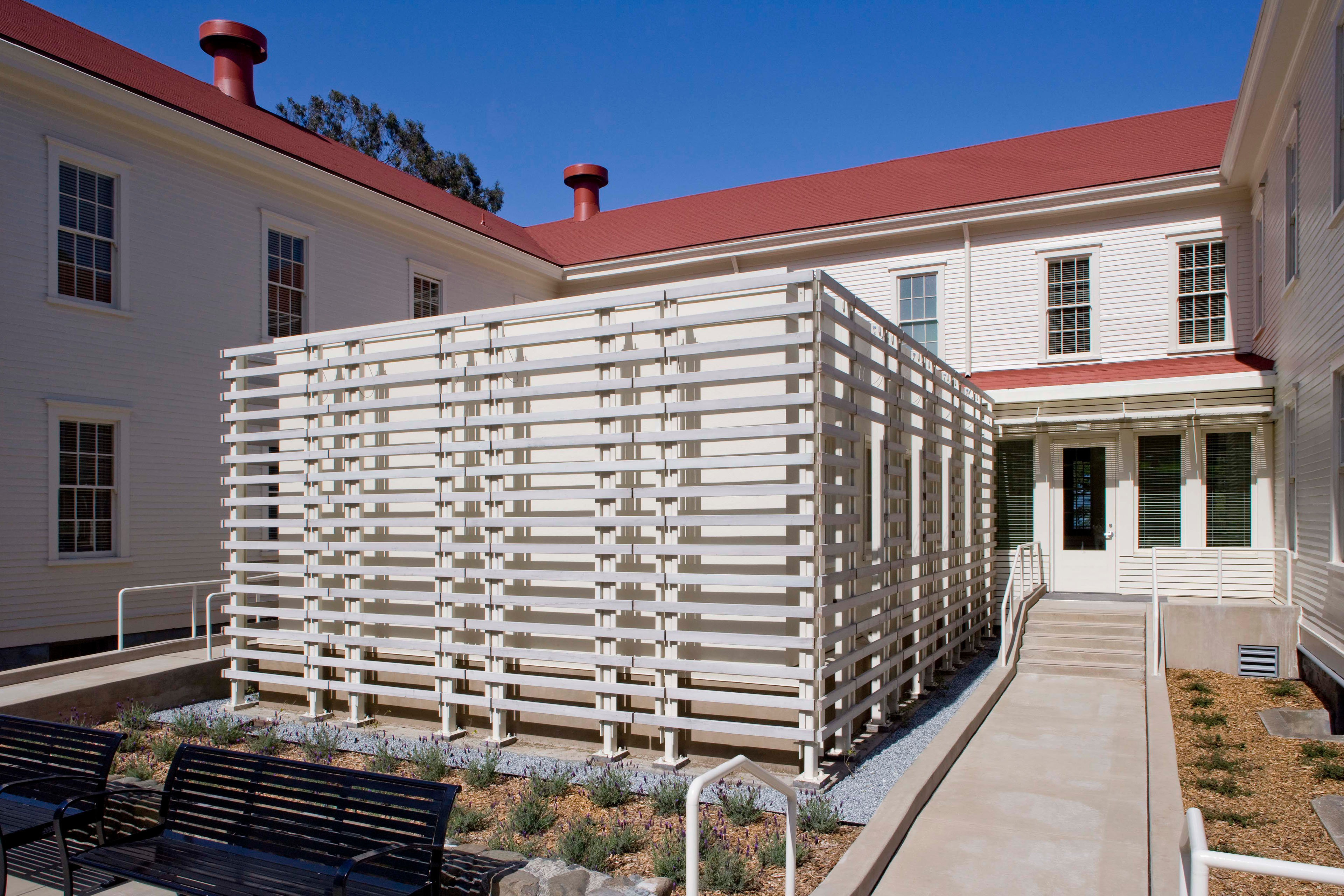 Courtyard of a historic white building with red roofs, accessible ramps, benches, and a large white slatted enclosure.