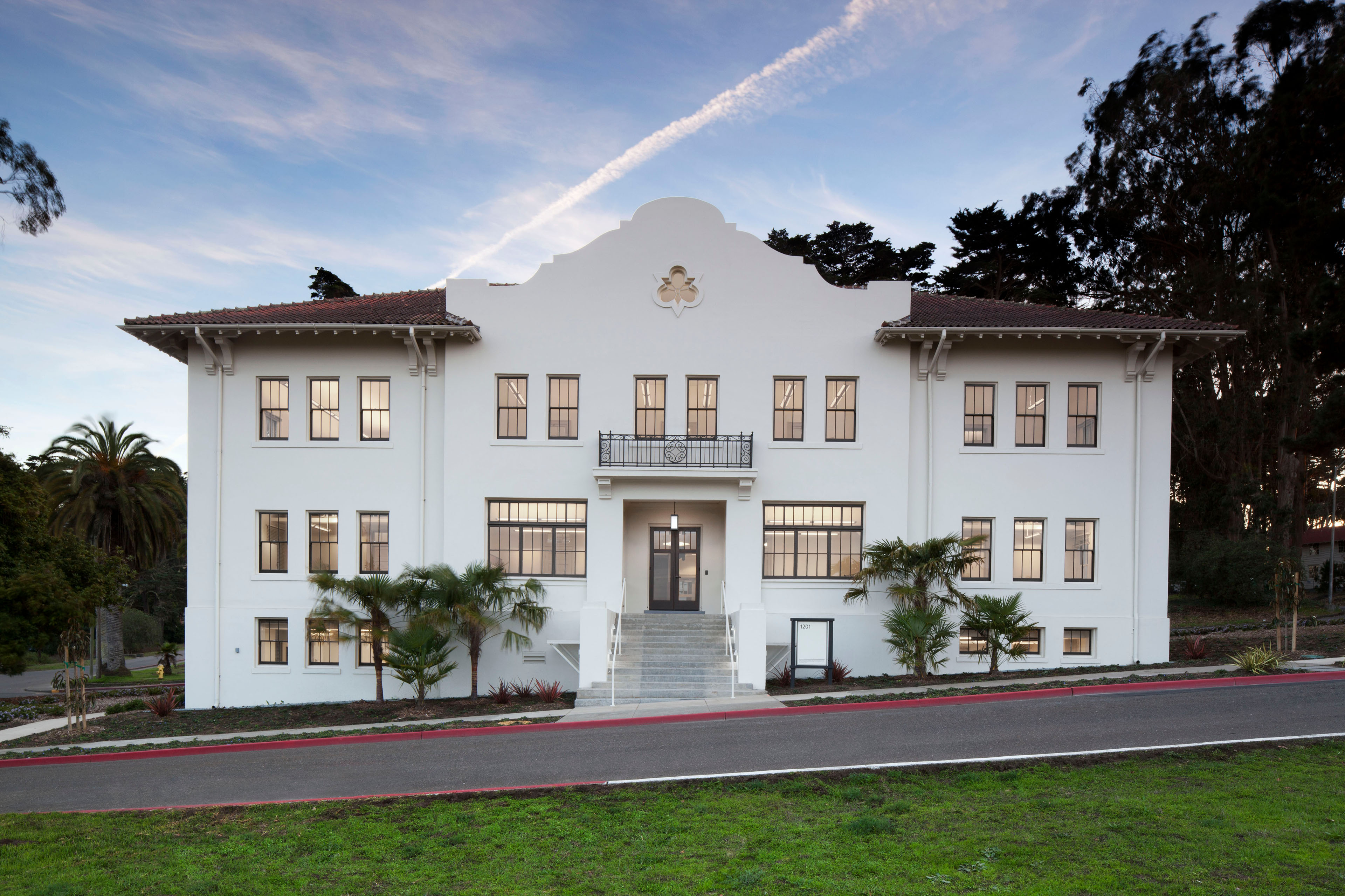 Front view of a symmetrical three-story historic building with white stucco walls, red tile roofs, dark-framed windows, and a central entrance reached by broad steps. A small balcony sits above the doorway, with landscaped palms, a road, and lawn in the foreground.