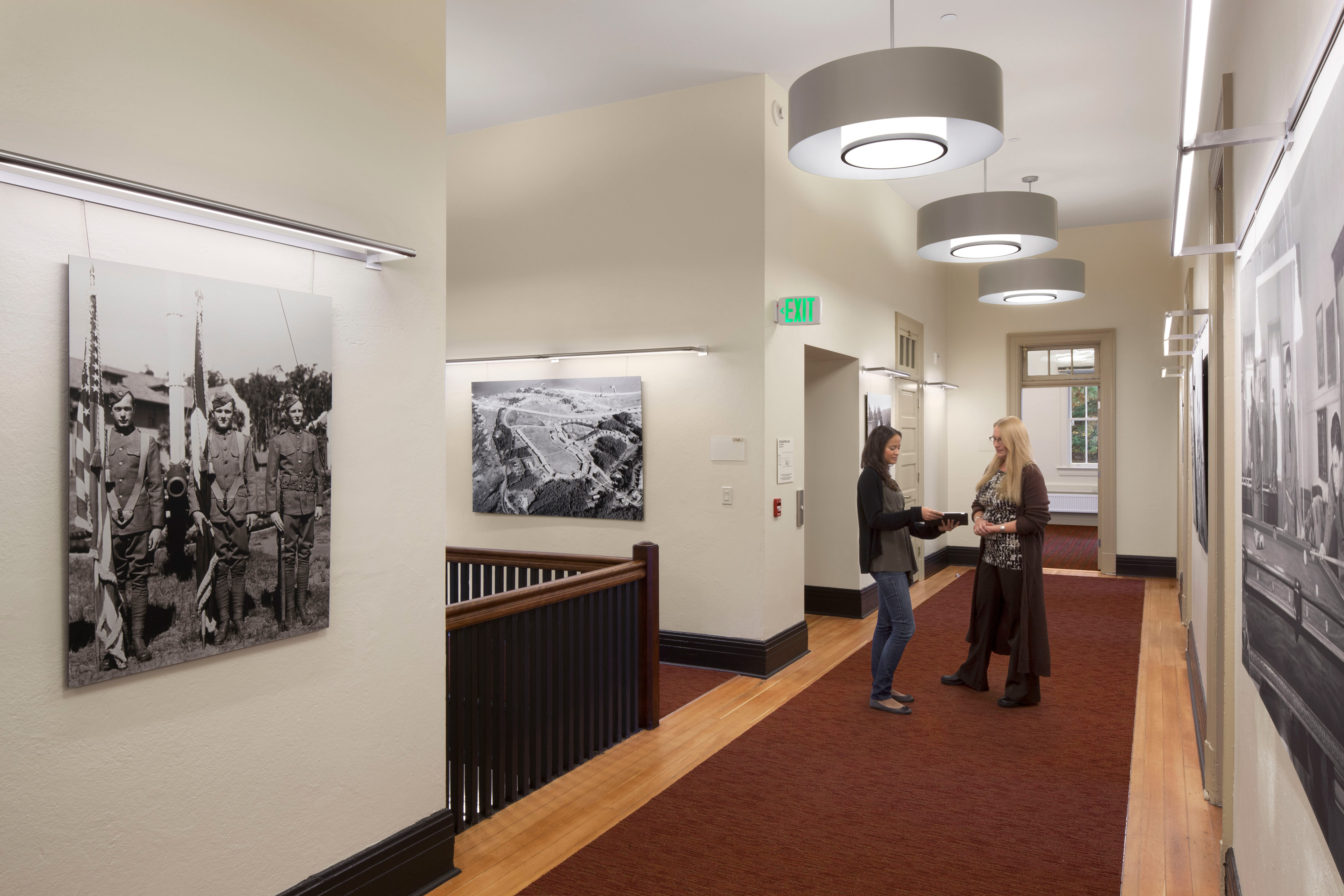 Renovated hallway displaying large black-and-white historical photographs. Two women talk beneath circular pendant lights, with wood flooring, a red carpet runner, and an illuminated exit sign.