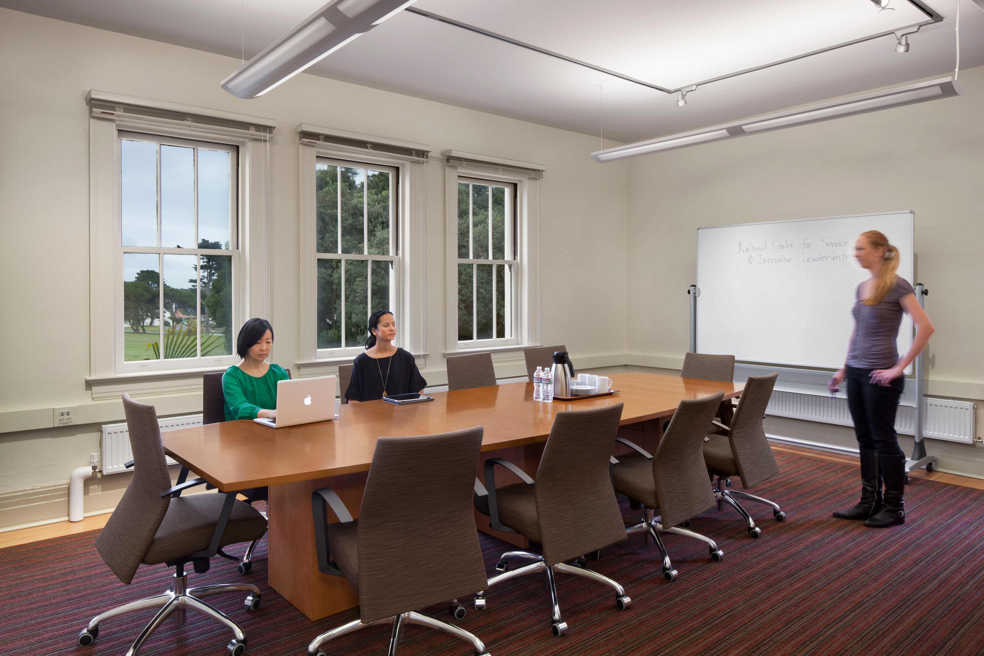 Conference room with a long wooden table and rolling chairs beneath suspended linear lights. Two women sit near large windows, one using a laptop, while another woman stands beside a whiteboard at the front of the room.