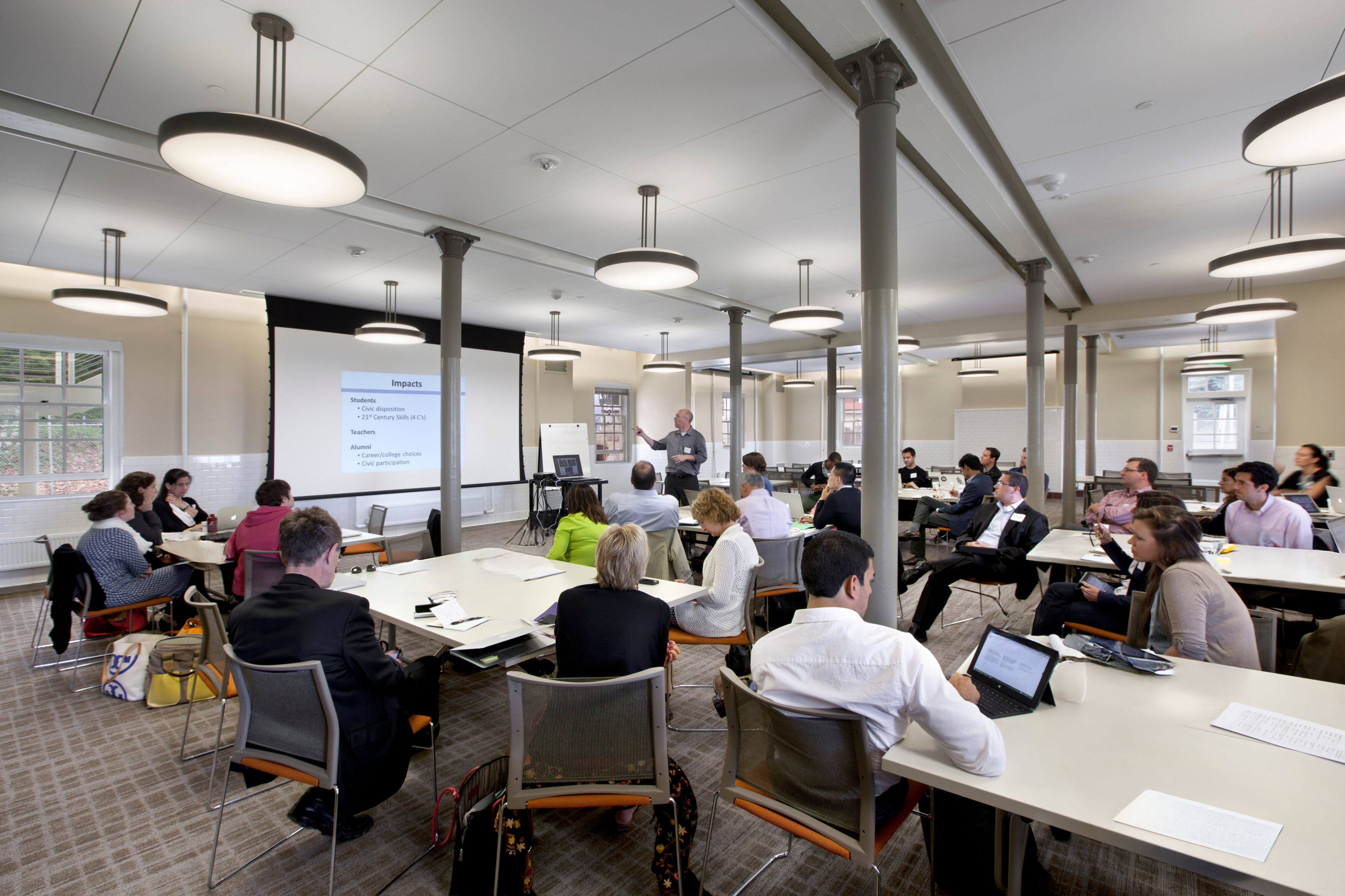 Large training room with attendees seated at tables facing a presenter and projected slide. Round pendant lights and gray columns span the renovated space, while participants take notes and use laptops.