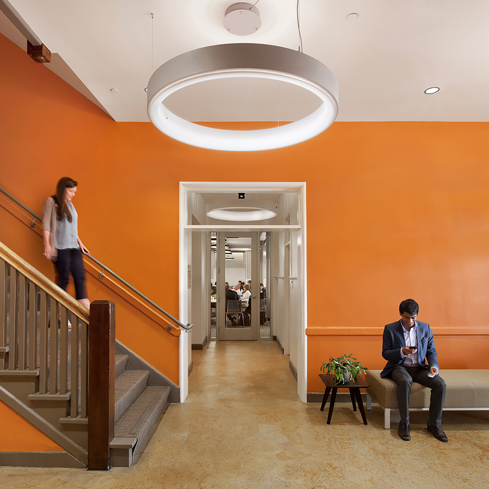 Interior landing with vivid orange walls, a carpeted staircase, and a large circular pendant light. A woman descends the stairs while a man sits on a bench looking at his phone; a meeting is visible through the doorway beyond.