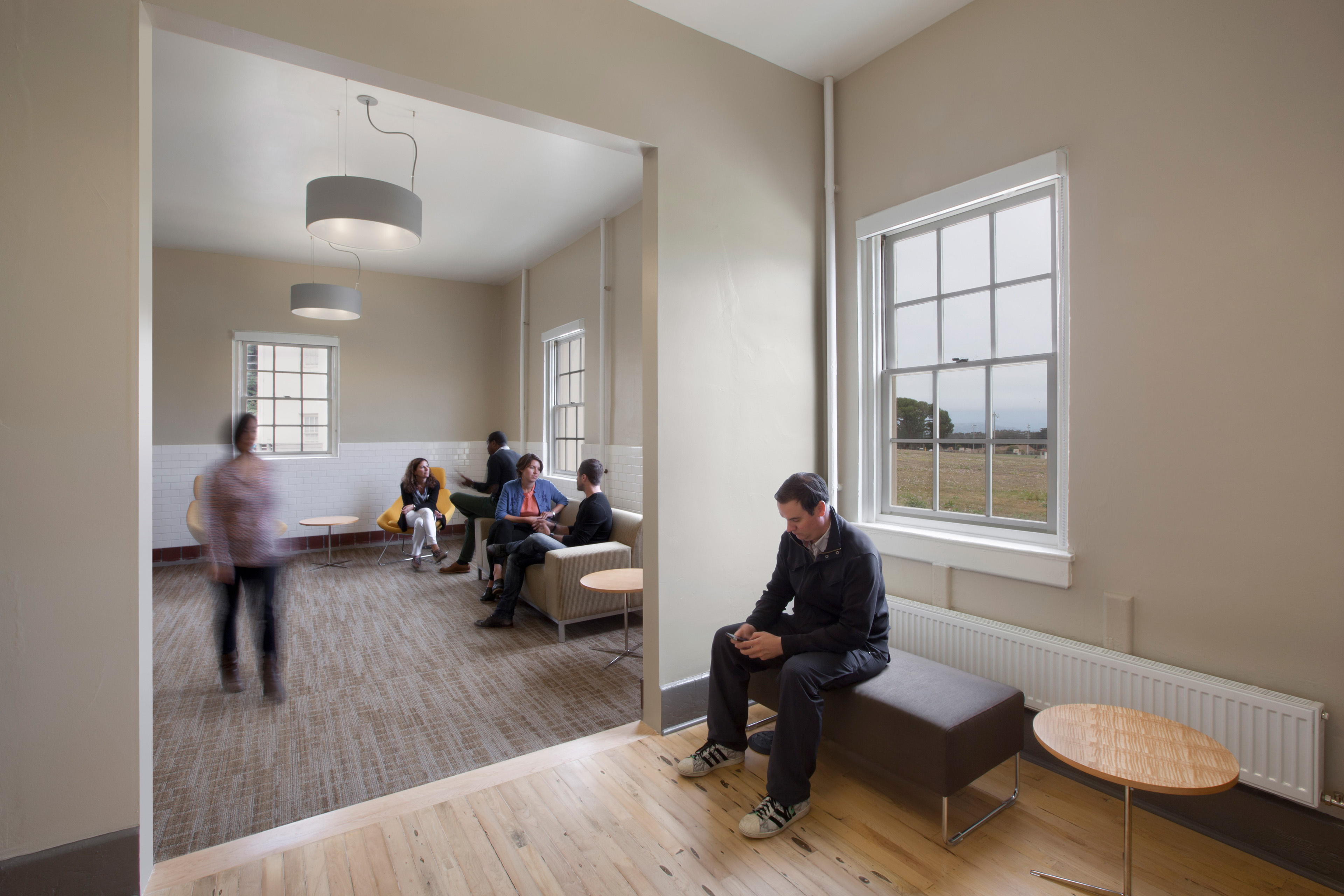 Interior lounge with neutral walls and large windows. A man sits on a bench looking at his phone in the foreground, while several people converse in an adjoining seating area.