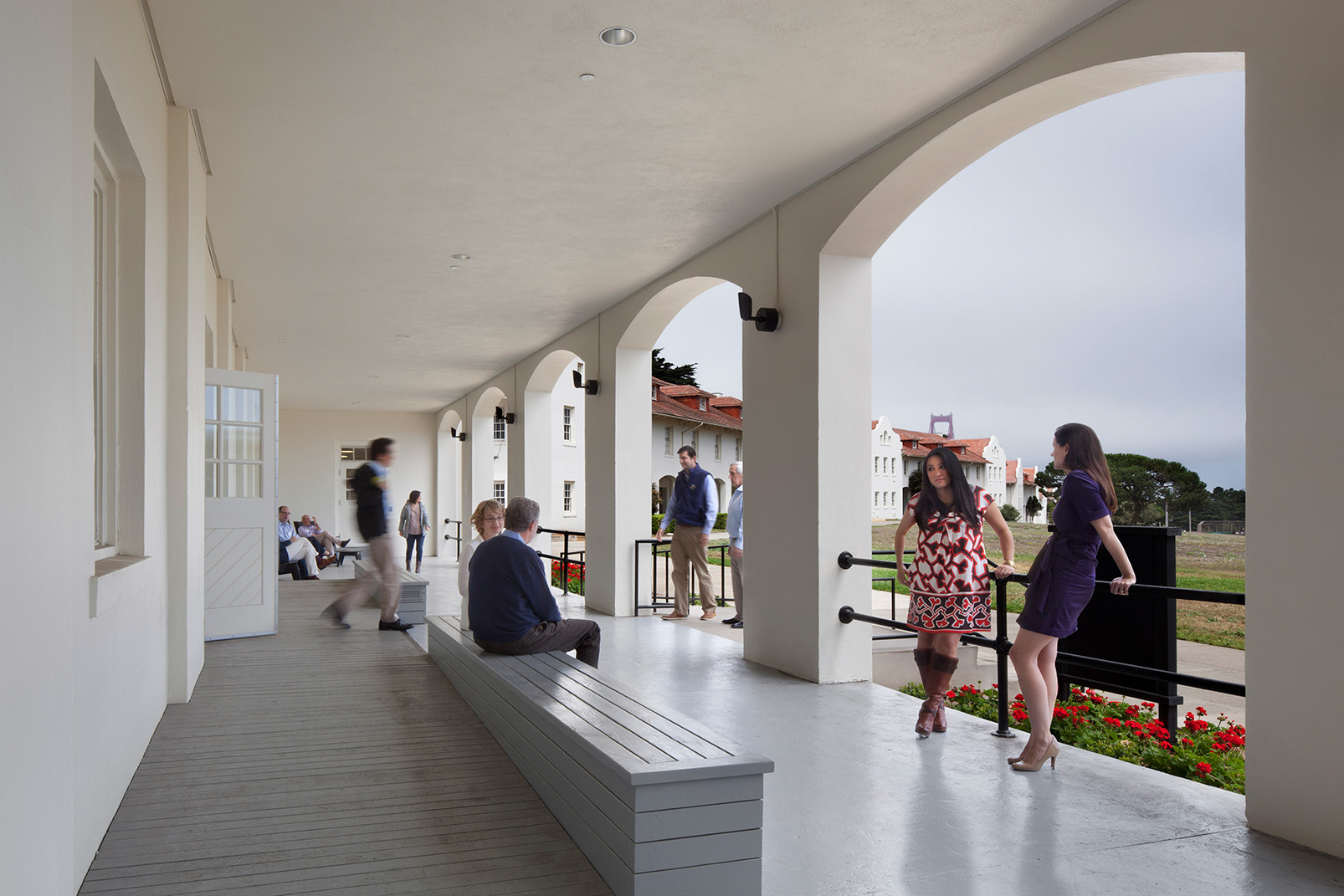 Covered exterior corridor with large white arches overlooking a lawn and neighboring red-roofed buildings. Several people sit, walk, or talk along the porch, while the Golden Gate Bridge is faintly visible through the mist in the distance.