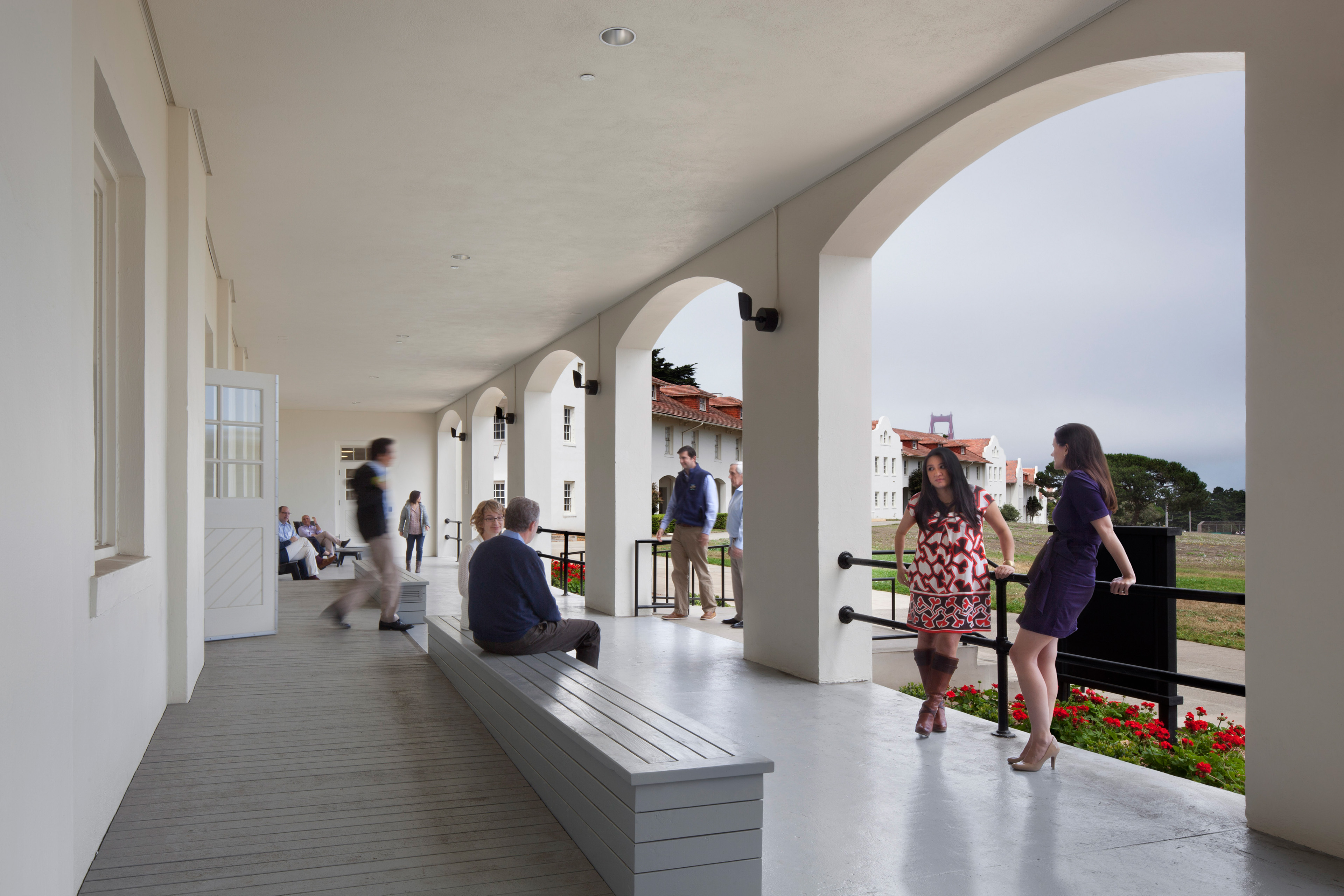 Covered exterior corridor with large white arches overlooking a lawn and neighboring red-roofed buildings. Several people sit, walk, or talk along the porch, while the Golden Gate Bridge is faintly visible through the mist in the distance.