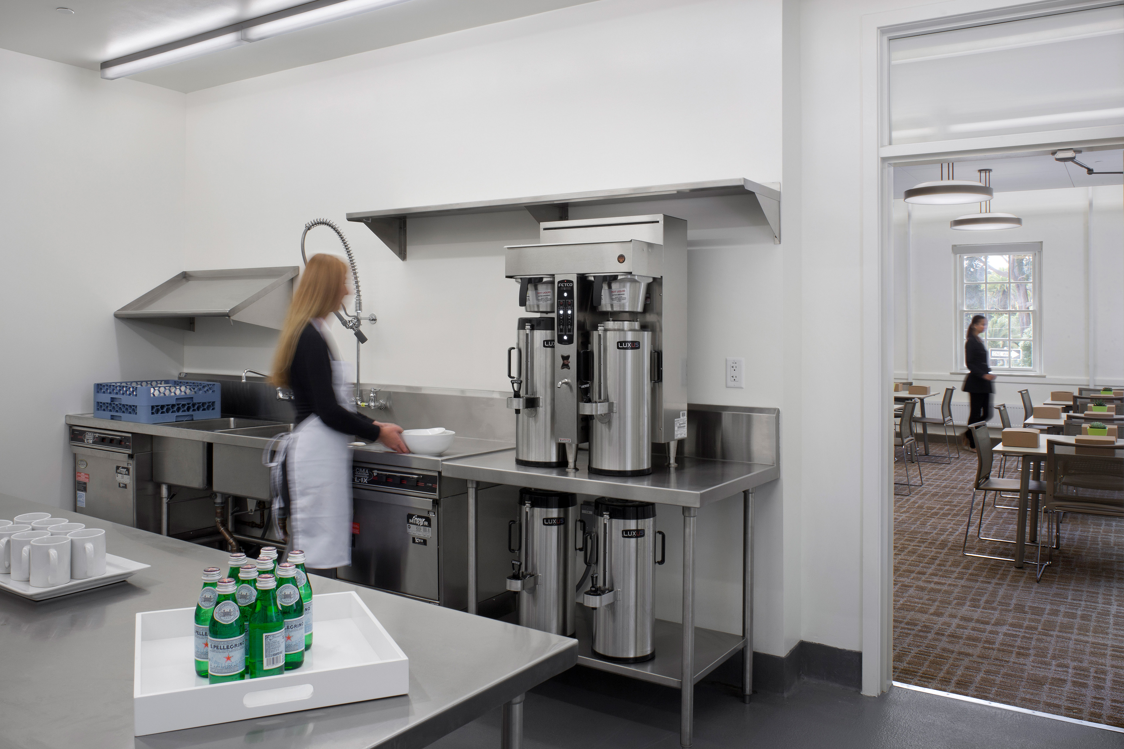 Commercial service kitchen with stainless steel counters, sinks, coffee urns, cups, and bottled water. A staff member works at the counter, with a furnished meeting room visible through the doorway.