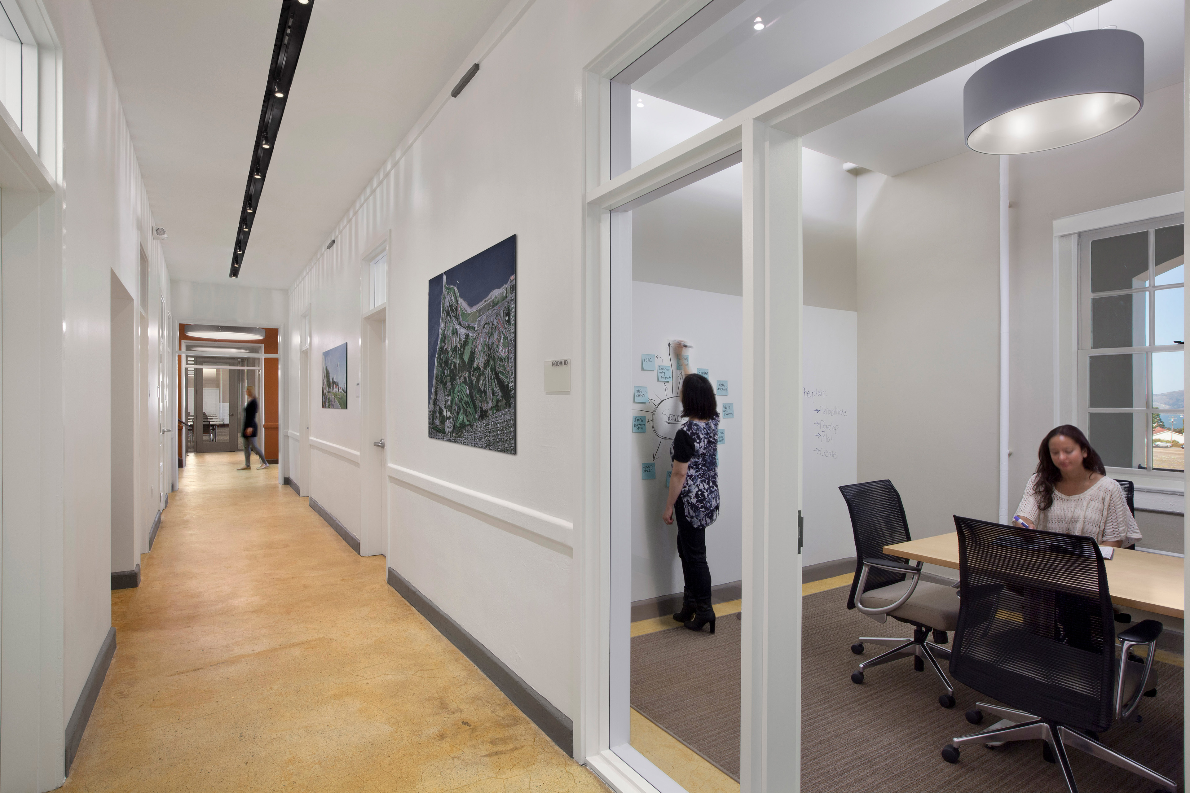 Bright office corridor beside a glass-walled meeting room. One woman writes on a wall covered with notes while another sits at the conference table, and a blurred person walks farther down the hallway.