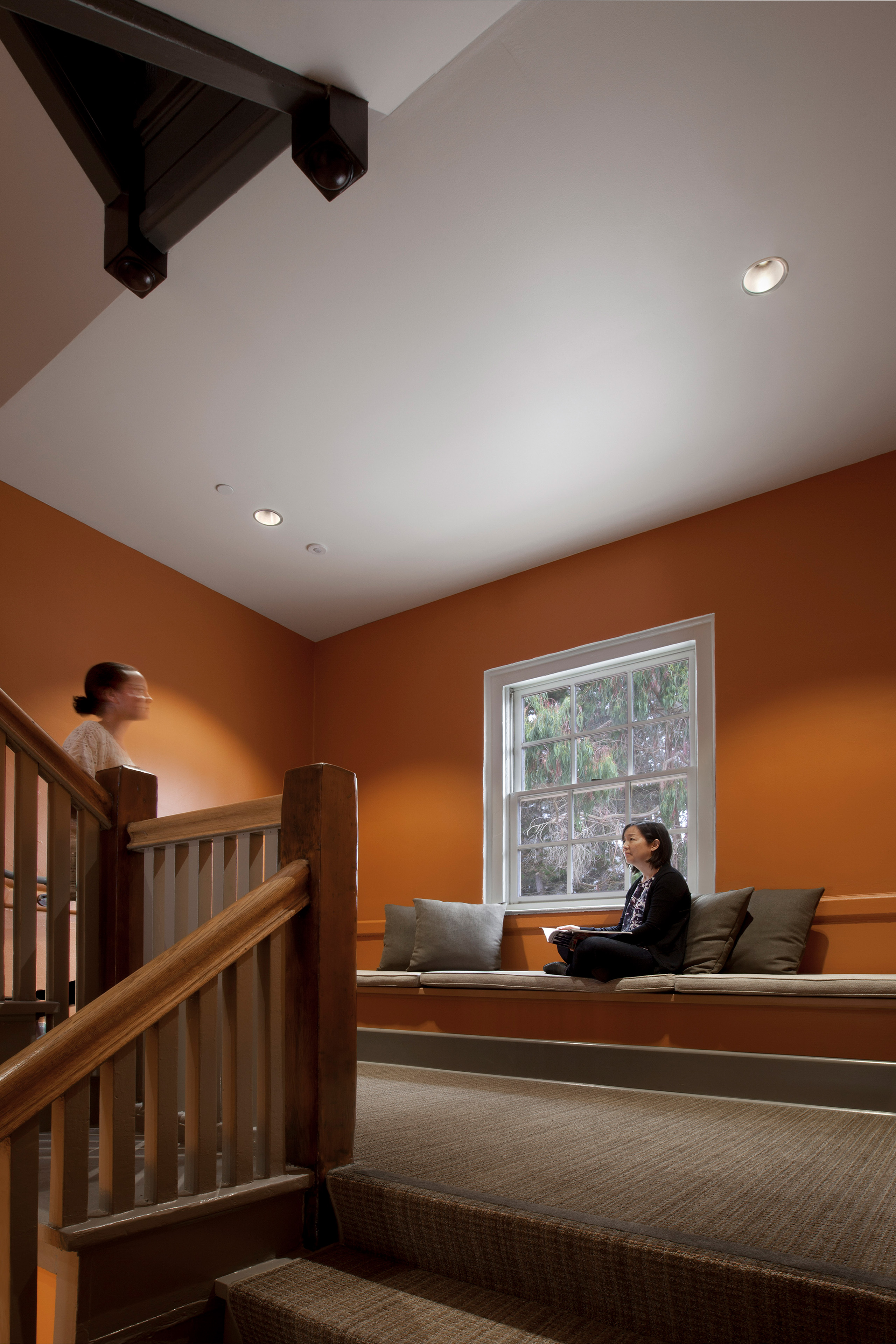 Interior stair landing with orange walls, exposed dark wood beams, and a broad window above a built-in cushioned bench. One woman sits reading on the bench while another, blurred in motion, walks up the wooden staircase.