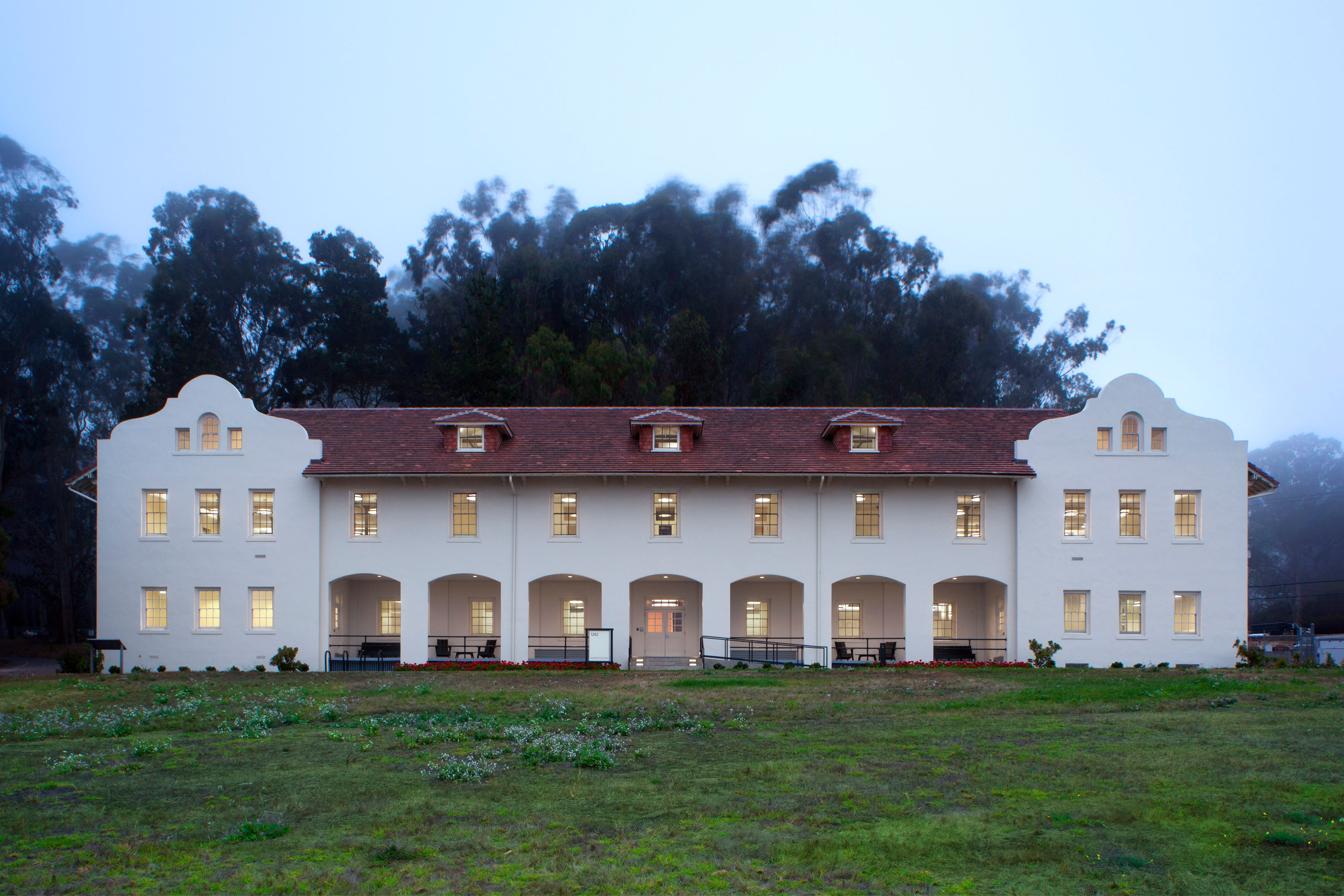 Front view of a symmetrical two-story historic building with white stucco walls, a red tile roof, arched ground-floor porches, and illuminated windows. A broad grassy lawn fills the foreground, with tall trees and light fog behind the building.