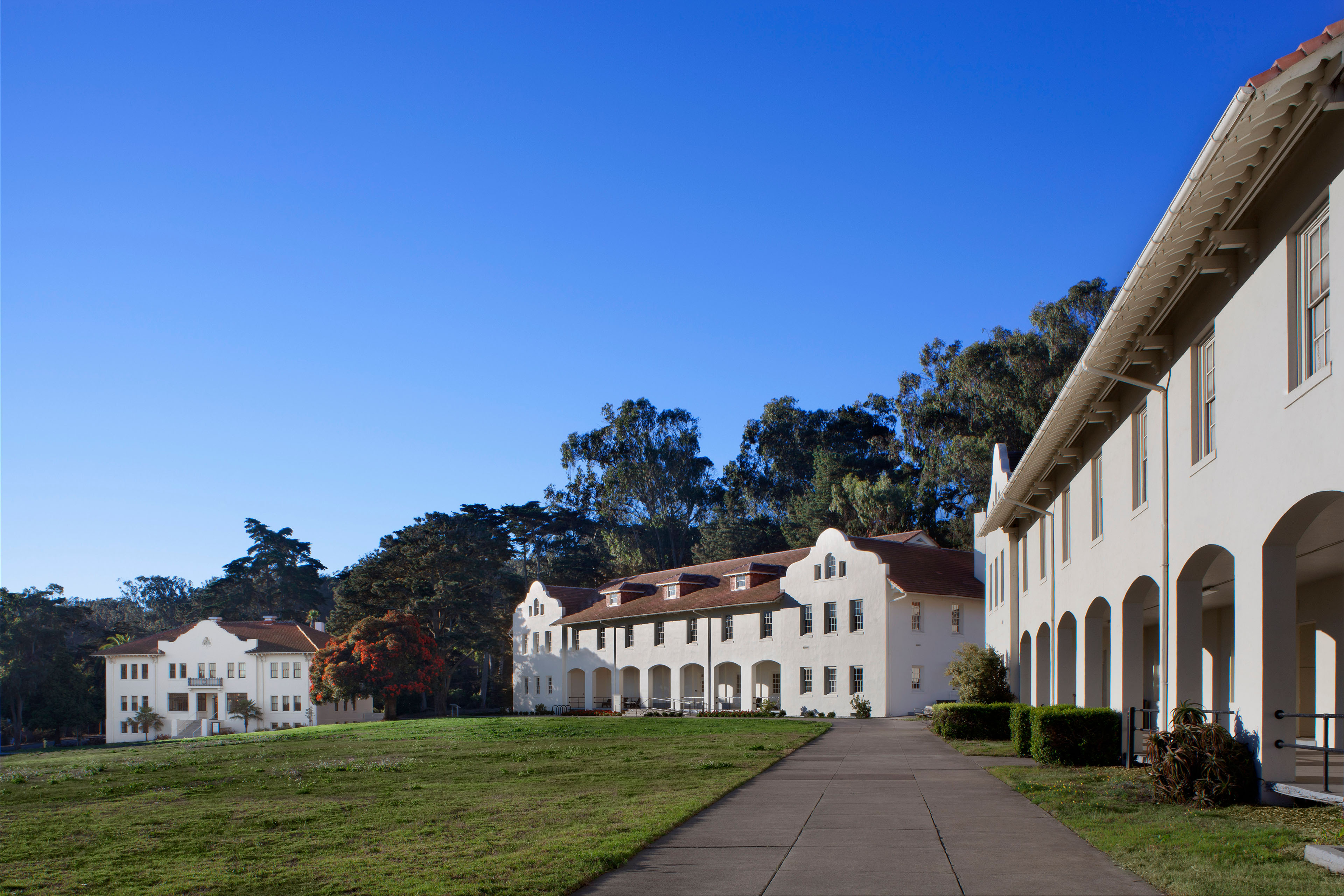 Wide exterior view of white historic buildings with red tile roofs and arched ground-floor porches arranged around a grassy courtyard. A paved walkway leads between the buildings, with mature trees behind them under a clear blue sky.