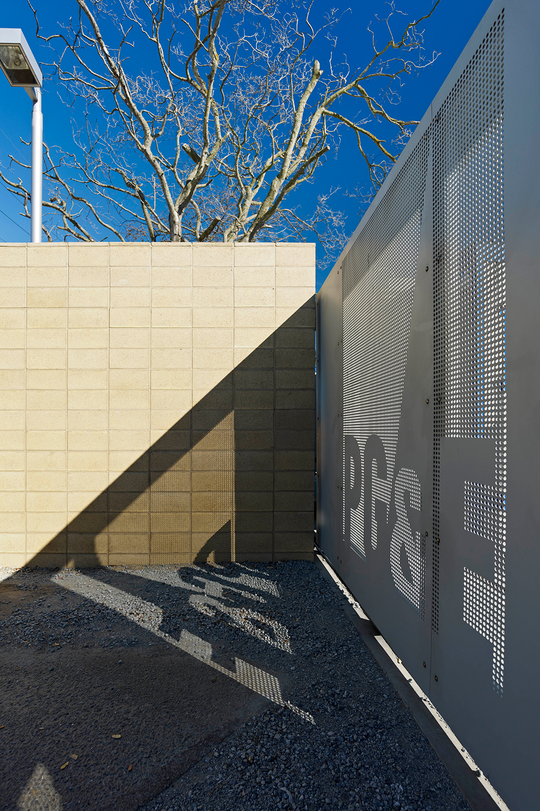 Architectural detail of a perforated metal screen beside a concrete block wall, casting patterned shadows across the ground beneath leafless trees and a clear blue sky.