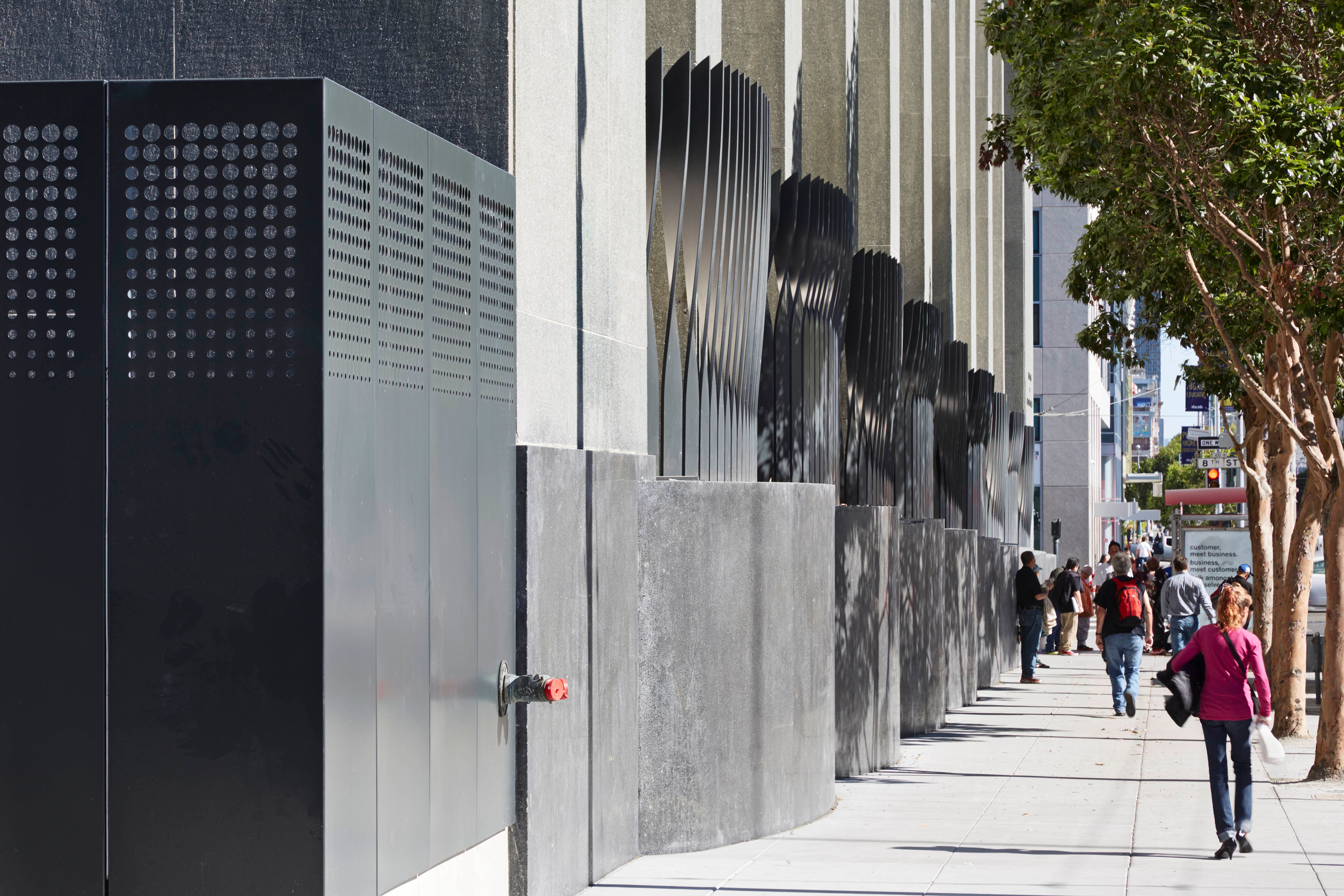 Street-level view of an urban utility building with perforated metal enclosures and sculptural vertical fins along the façade, with pedestrians and street trees lining the sidewalk.