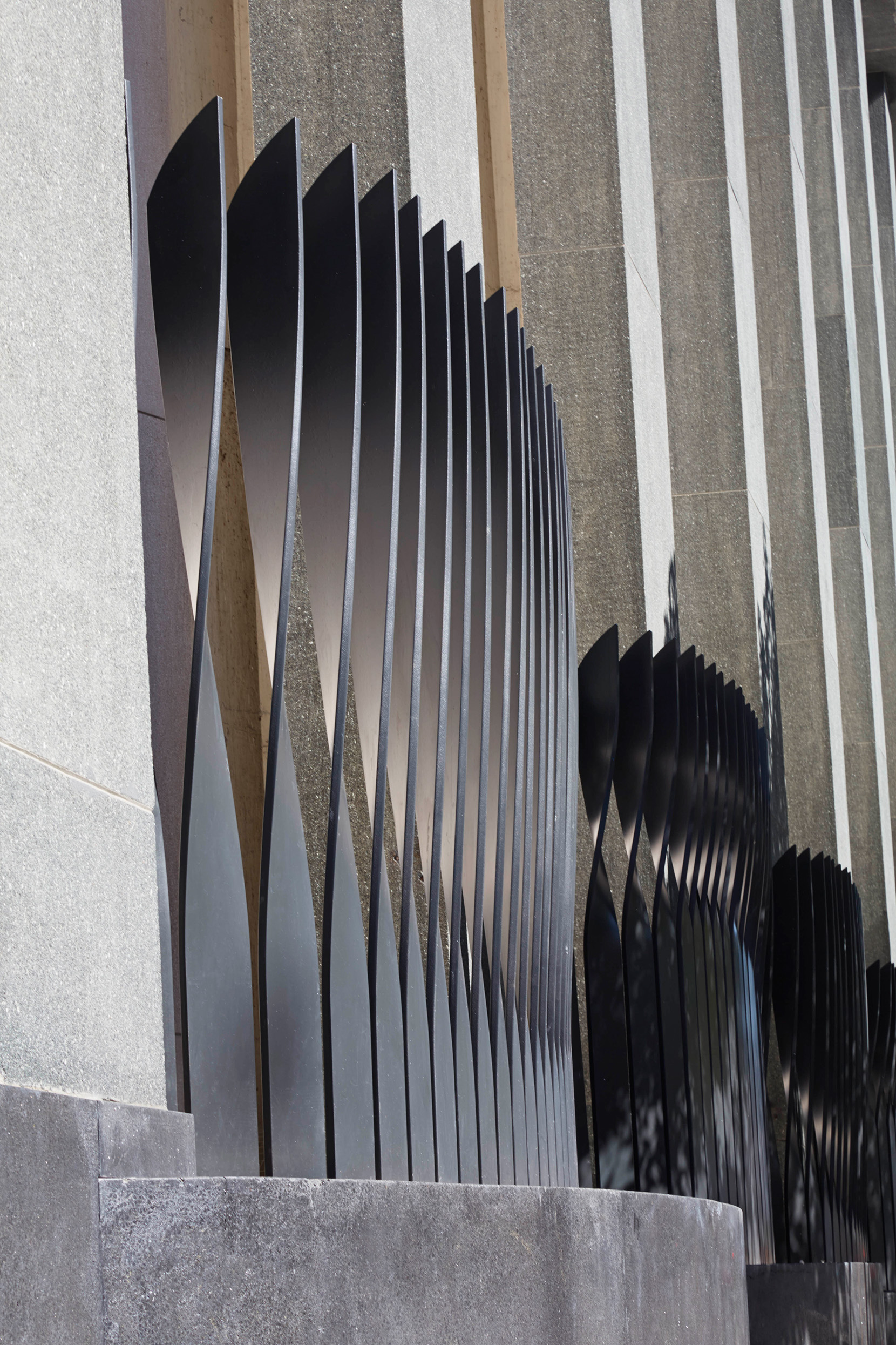 Close-up of sculptural black metal fins projecting from a textured concrete façade, creating a rhythmic pattern of light and shadow.