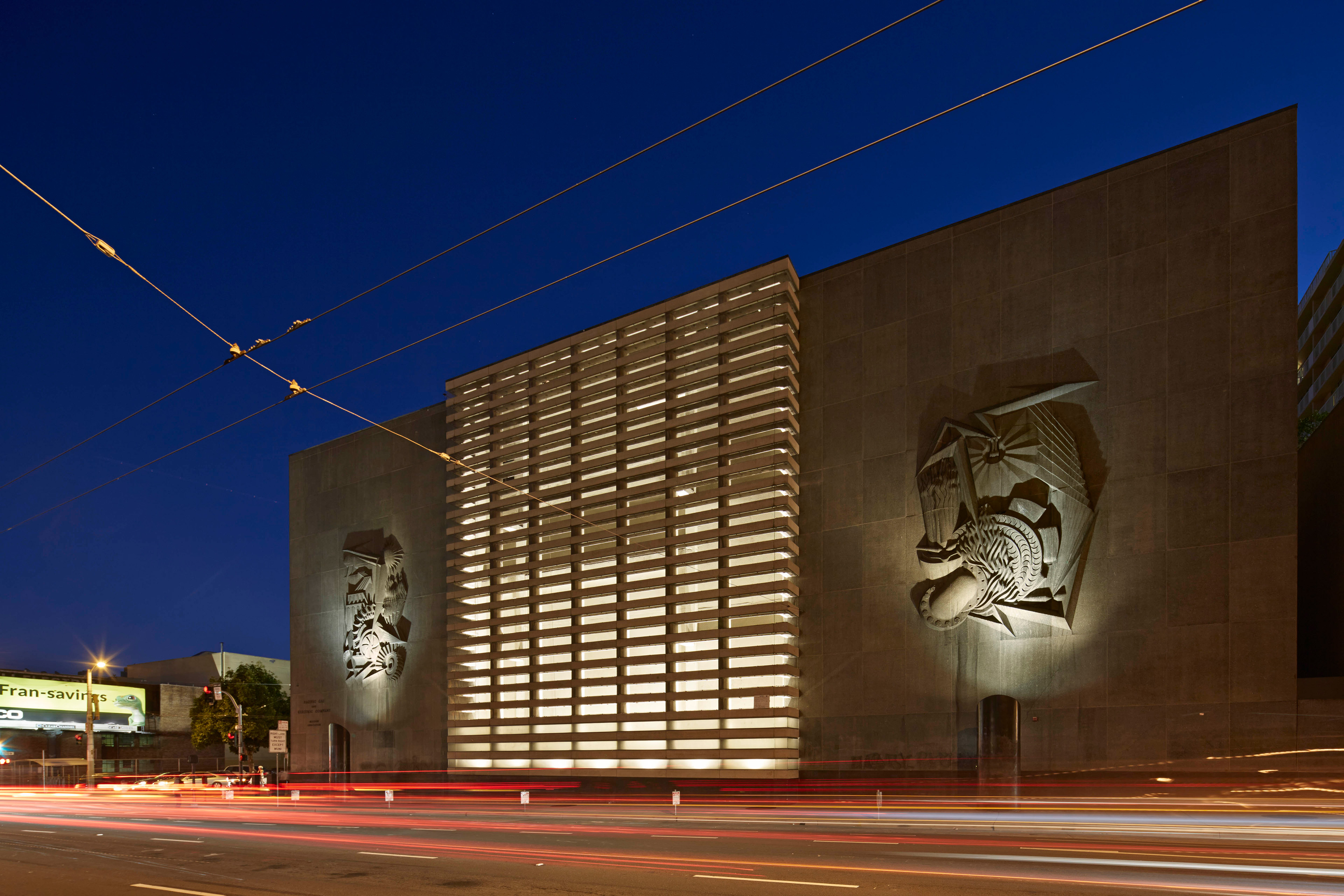 Illuminated urban utility building at night with monumental concrete walls, a glowing louvered central façade, sculptural reliefs, and traffic light trails across the street in the foreground.
