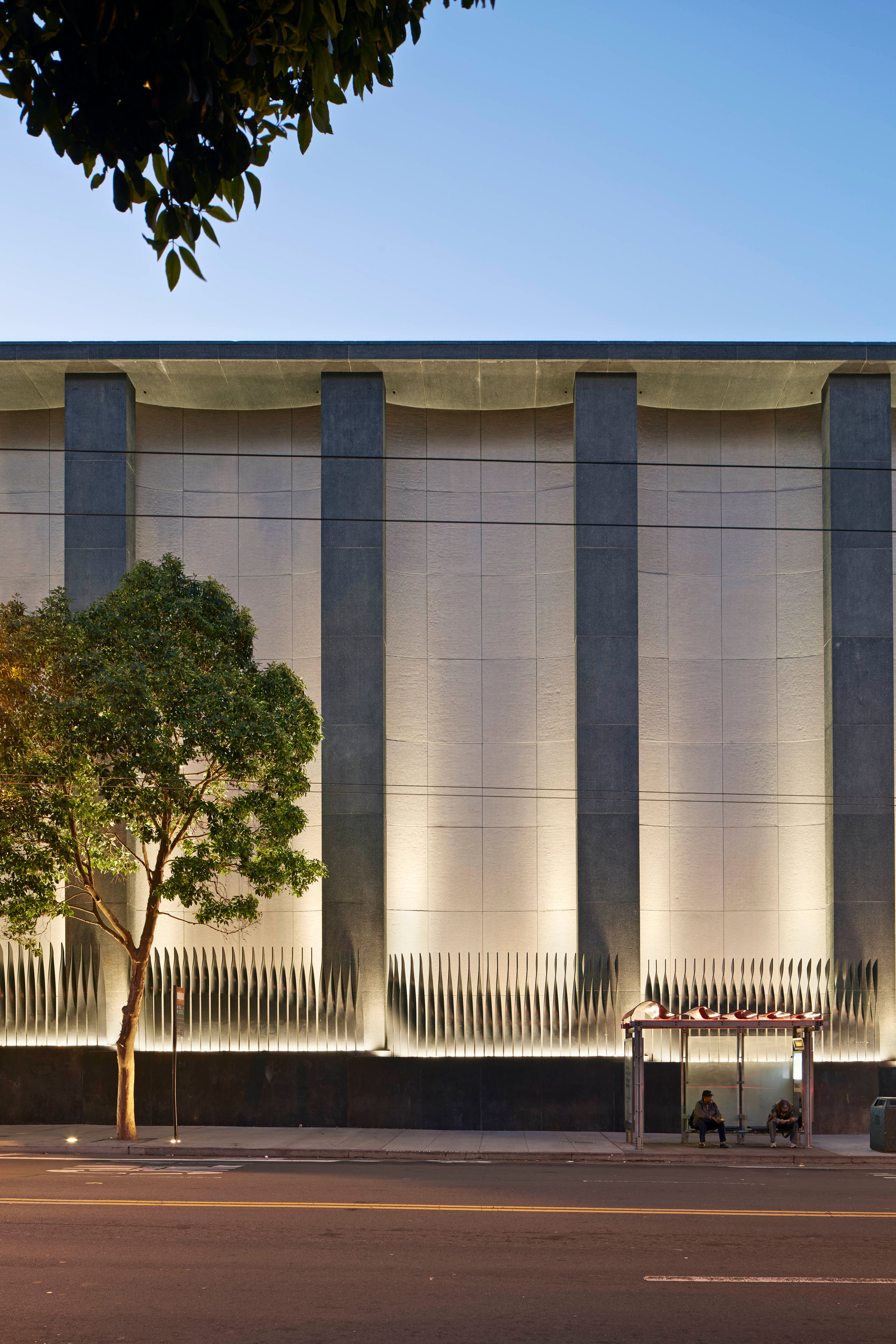 Street-facing utility building façade at dusk with tall concrete piers, illuminated metal screening at the base, a mature tree, and people waiting at a bus shelter.