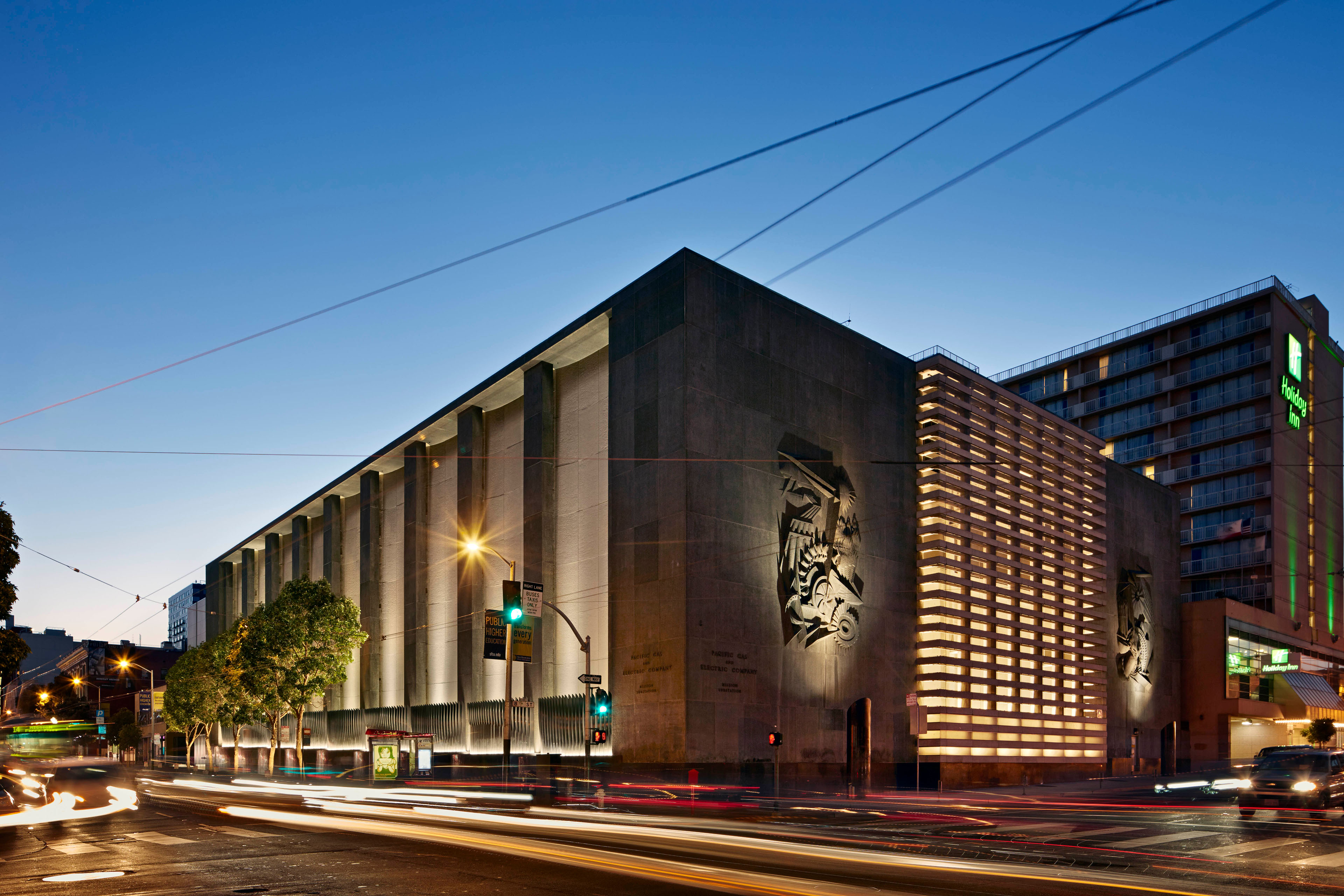 Large urban utility building at dusk with monumental concrete walls, illuminated vertical fins, sculptural reliefs, and glowing corner screening; traffic light trails animate the intersection in the foreground.