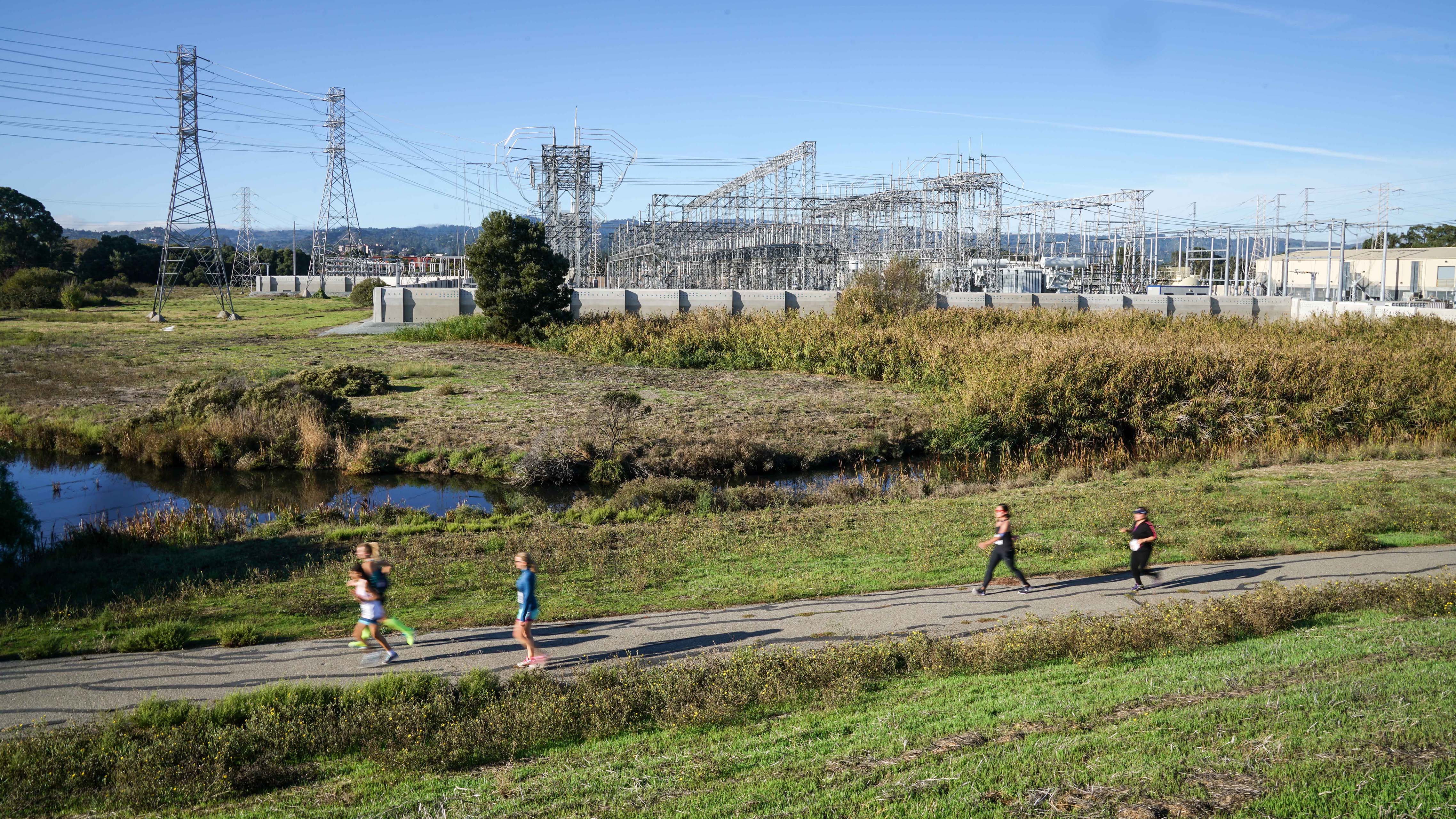 Wide view of an electrical substation beside a wetland and public trail, with runners and walkers in the foreground and transmission infrastructure extending across the landscape.