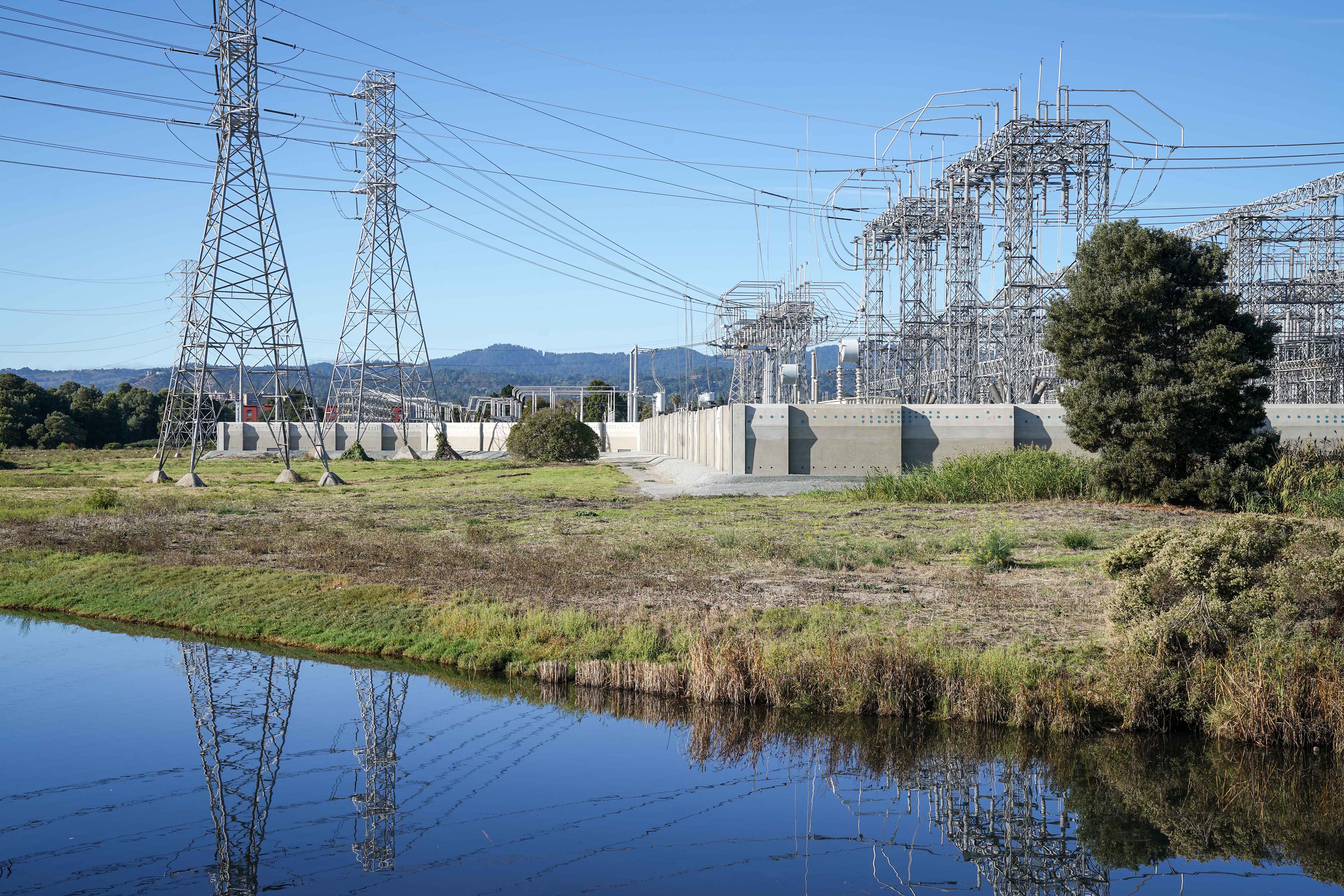 Electrical substation beside a wetland, with transmission towers, utility infrastructure, and a low precast concrete perimeter wall reflected in the water.