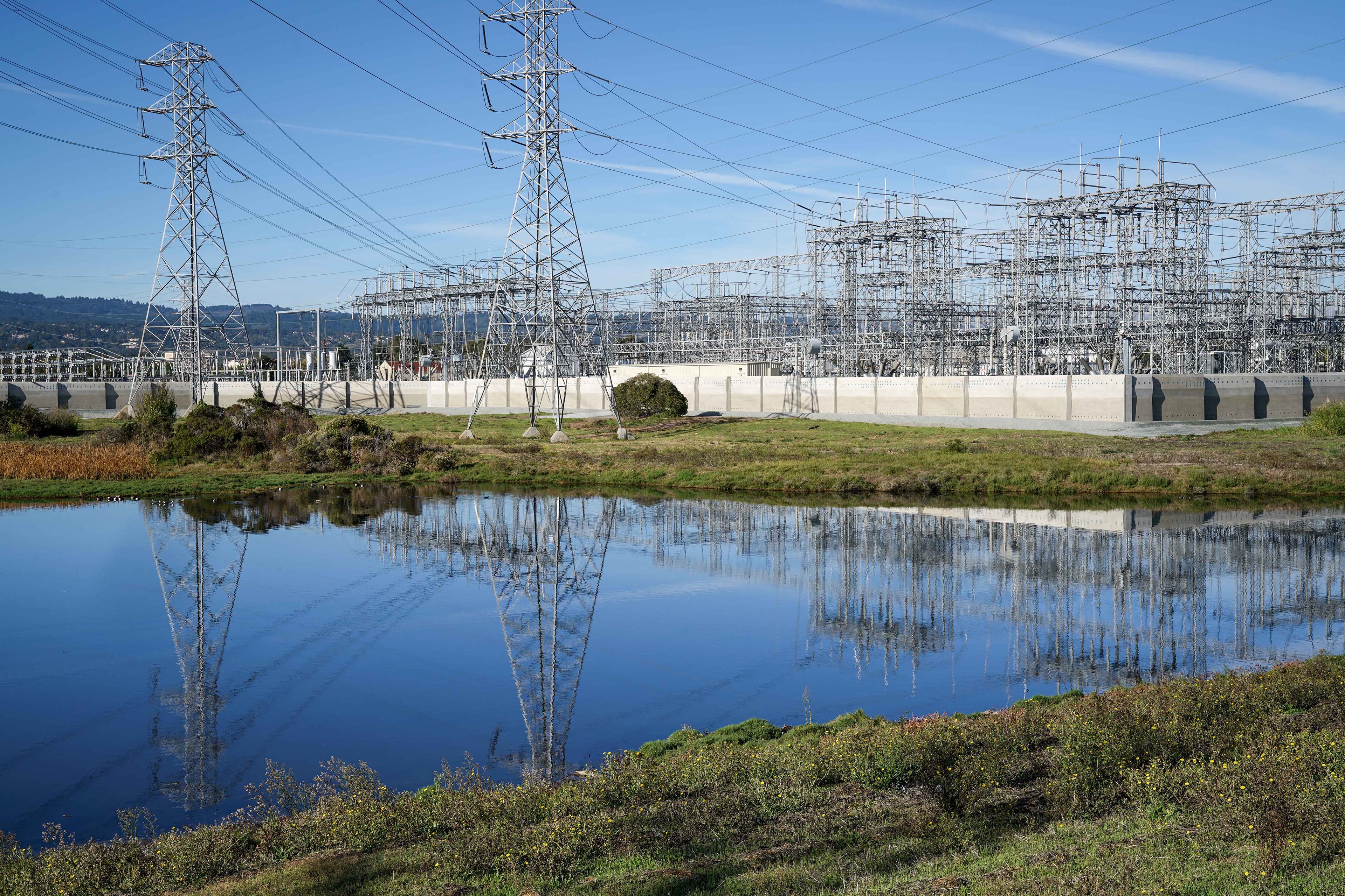 Electrical substation beside a wetland, with transmission towers and dense utility infrastructure reflected in the water and low hills in the distance.