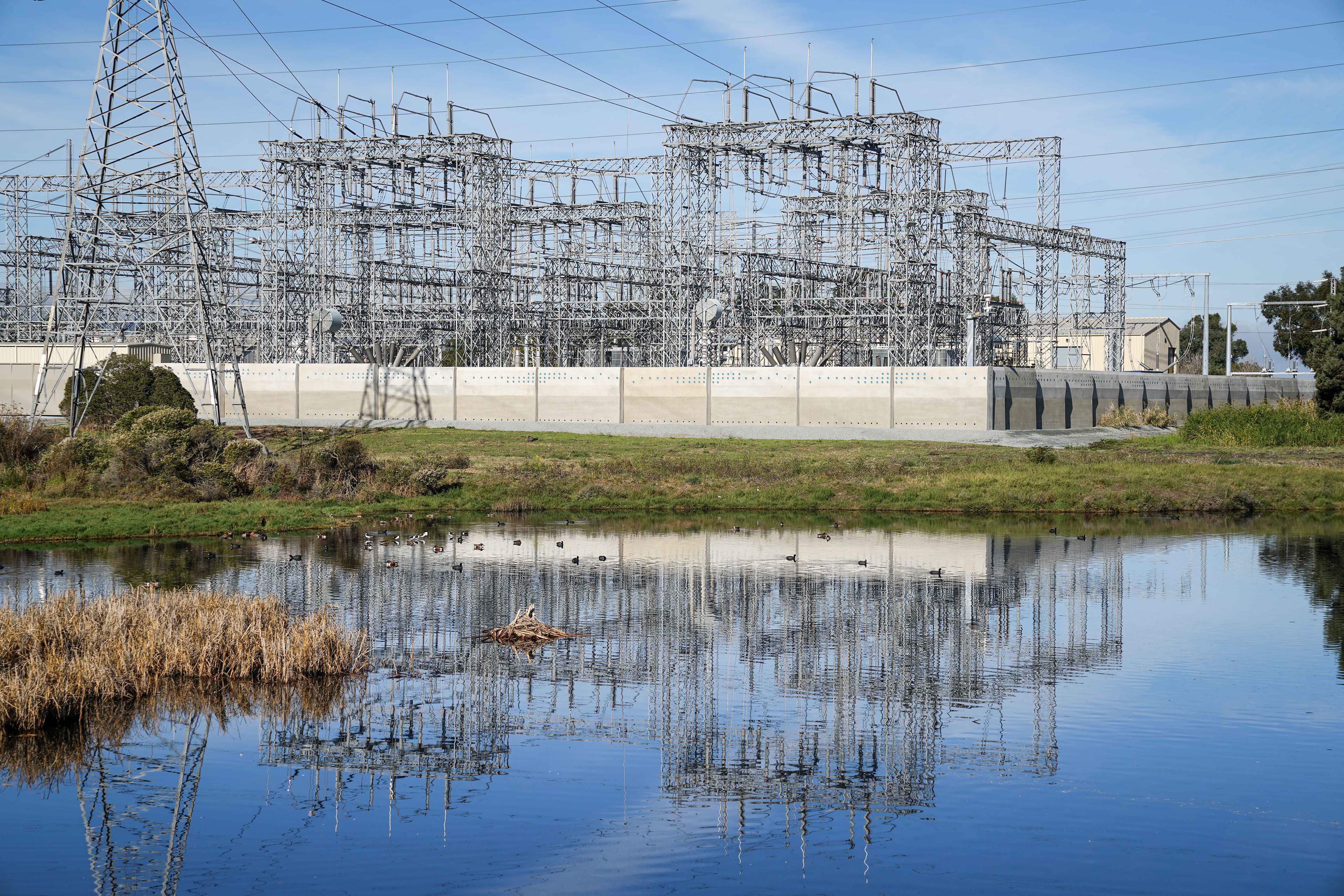 Electrical substation set beside a wetland, with dense steel transmission infrastructure rising behind a long precast concrete perimeter wall and reflected in the water.