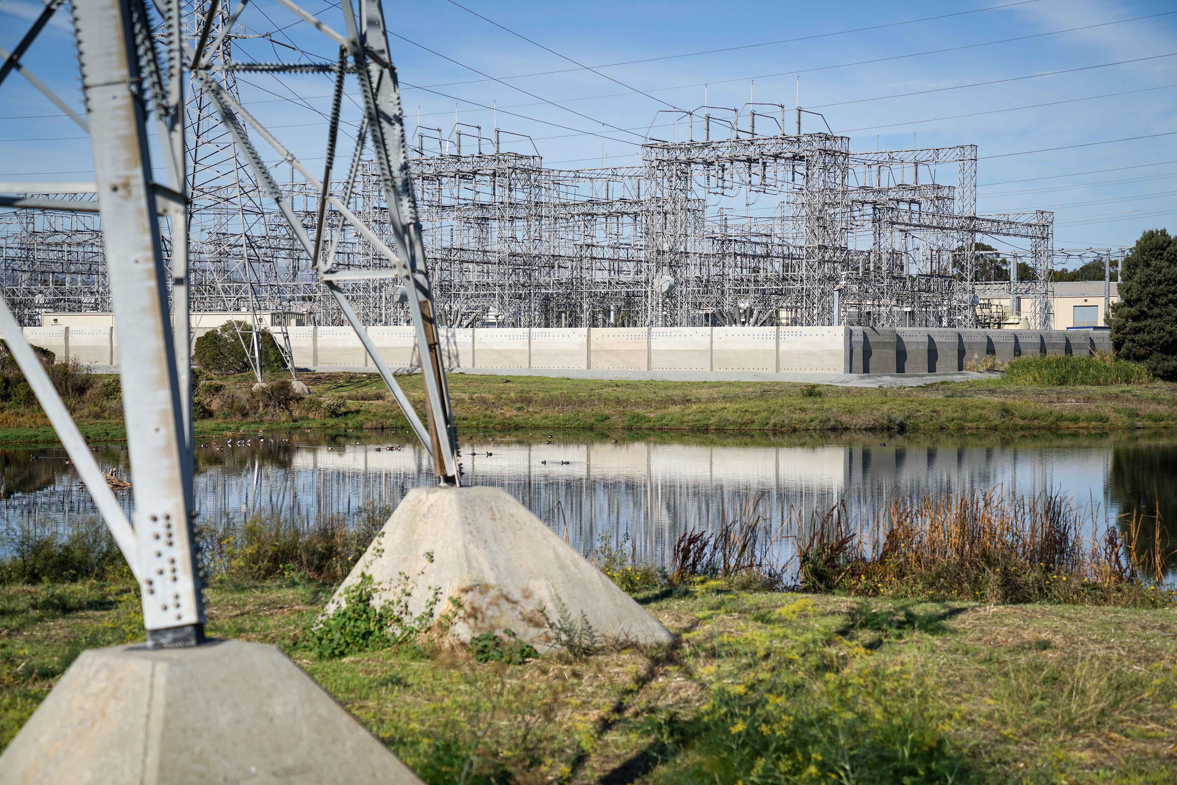 View across a wetland toward an electrical substation and precast perimeter wall, framed in the foreground by the base of a transmission tower and low native vegetation.