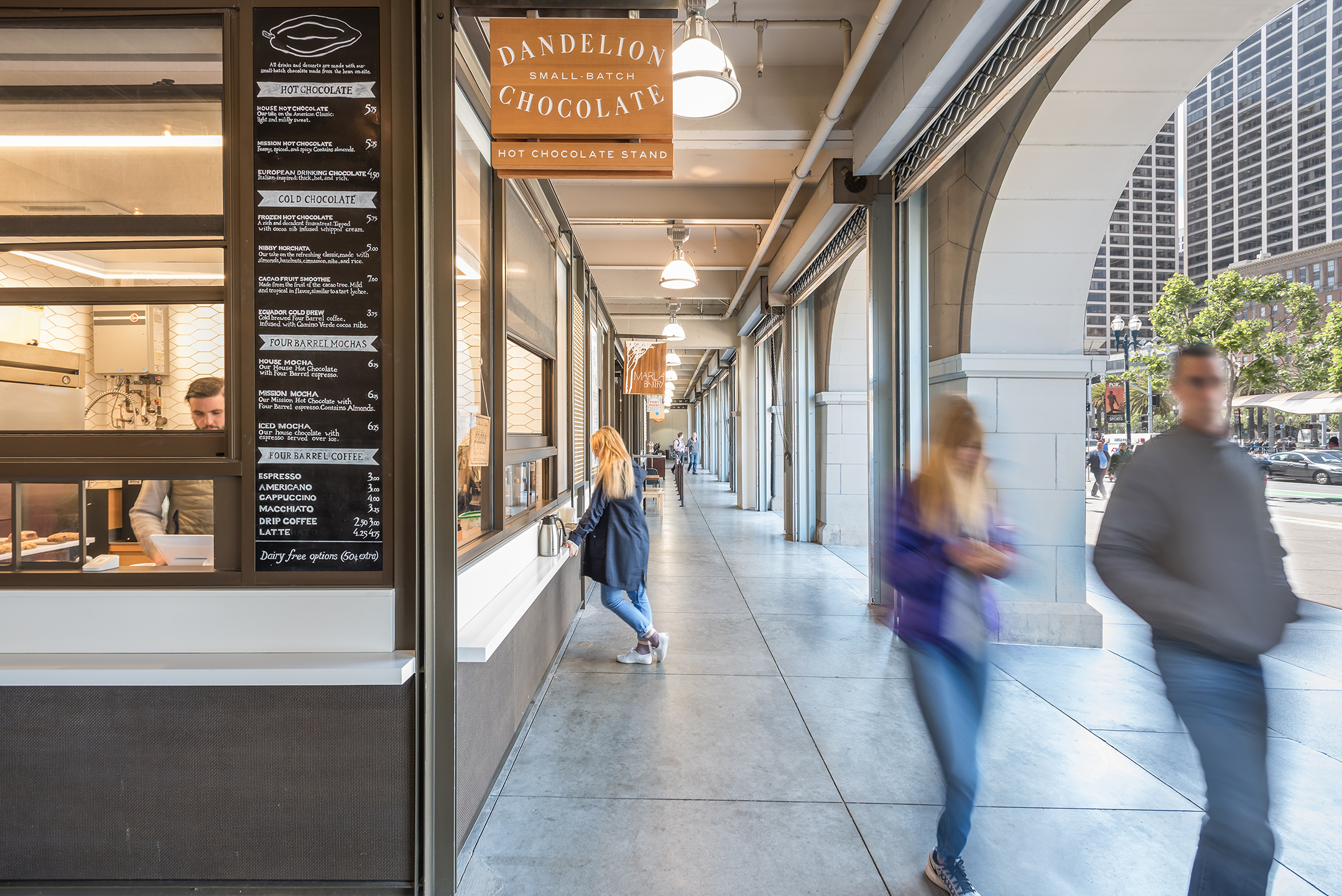 Dandelion Chocolate stand beneath a historic arched arcade, with a menu board, service counter, and pedestrians moving through the open marketplace corridor.
