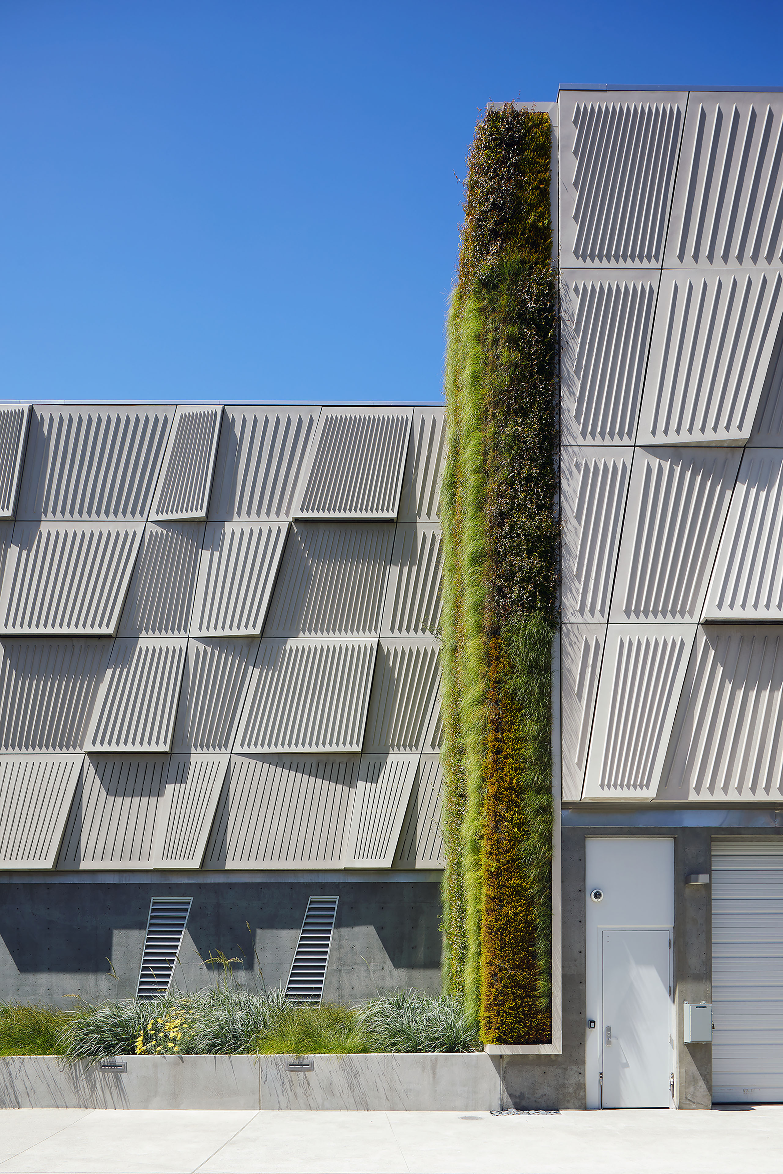 Close exterior view of a vertical living wall set between faceted metal façade panels, with concrete walls, planting beds, and service doors below.