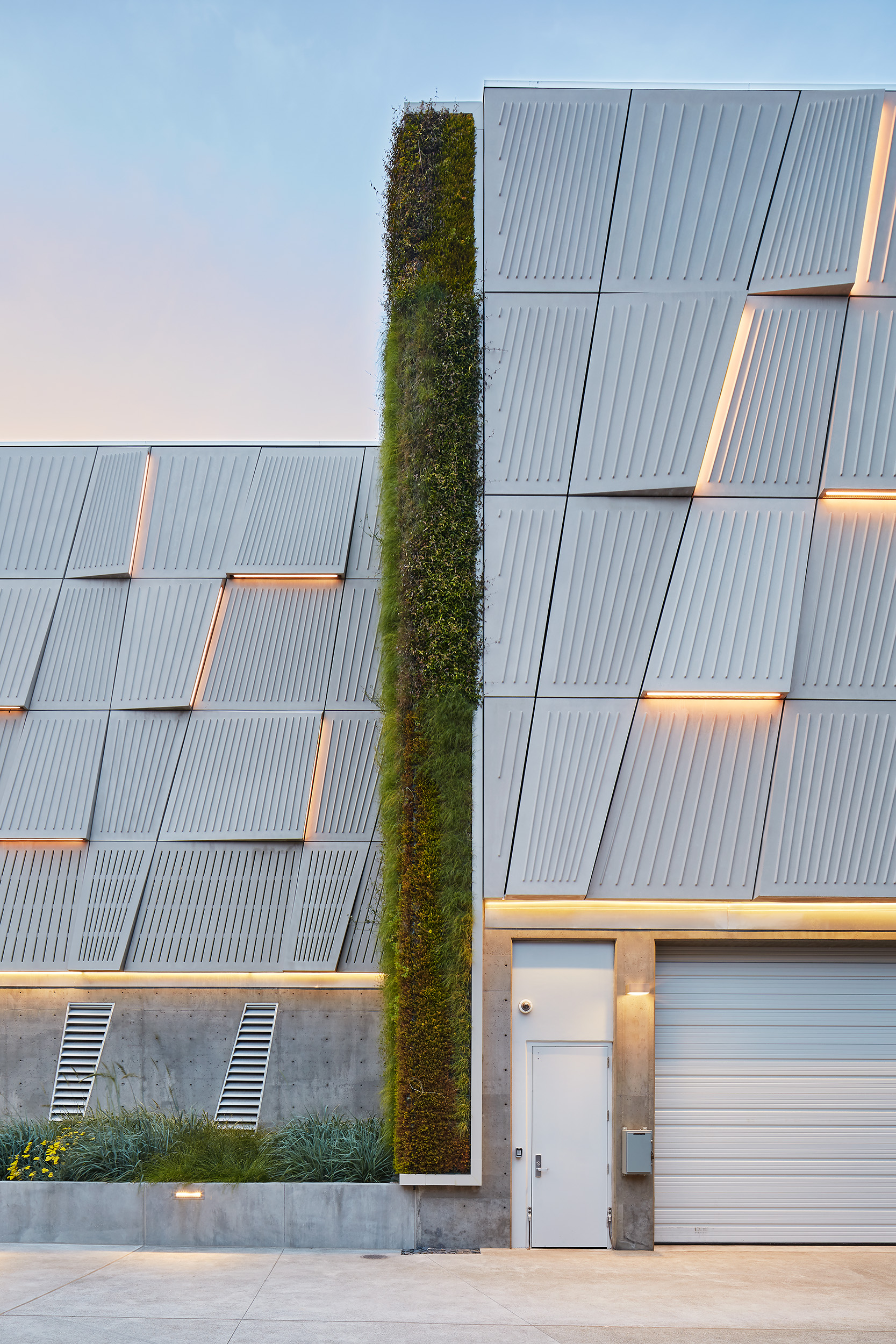 Tall vertical living wall framed by patterned metal façade panels with integrated lighting, above concrete service doors and landscaped planting beds.