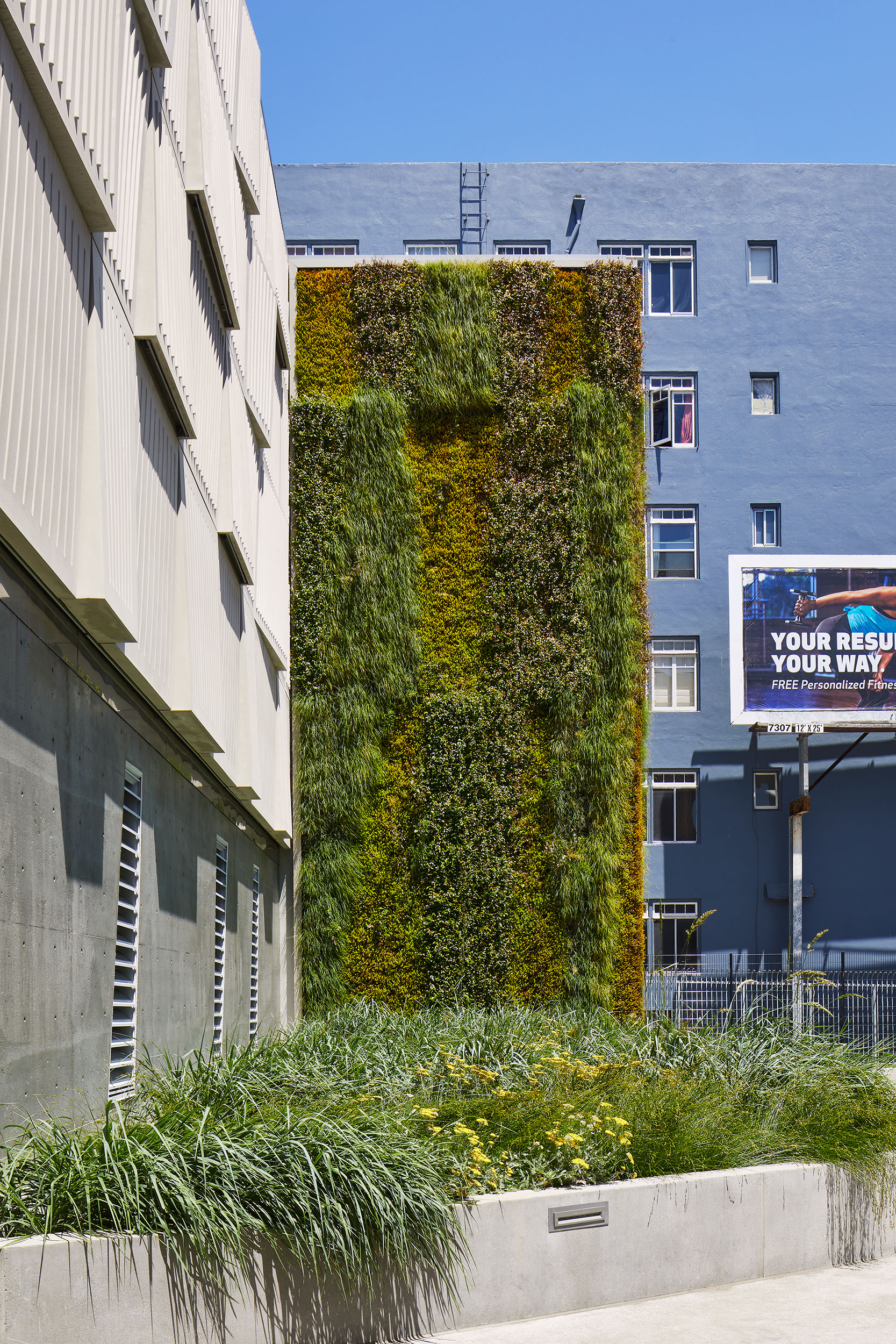 Tall living wall of varied green plantings integrated into a utility building façade, with exposed concrete, ventilation louvers, and a landscaped planter at the base.