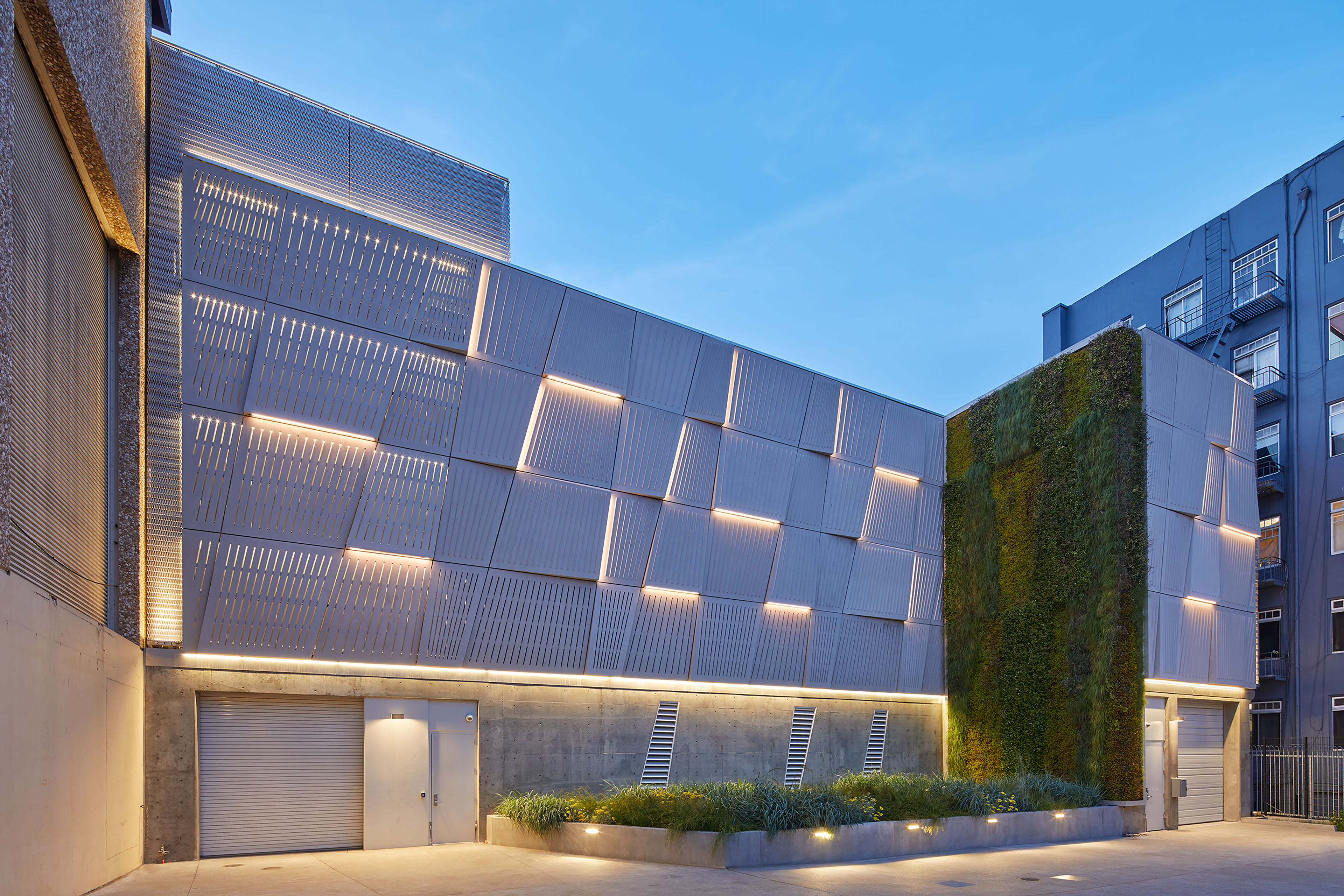 Utility building courtyard at dusk with patterned metal façade panels, integrated lighting, exposed concrete, landscaped planters, and a vertical living wall.