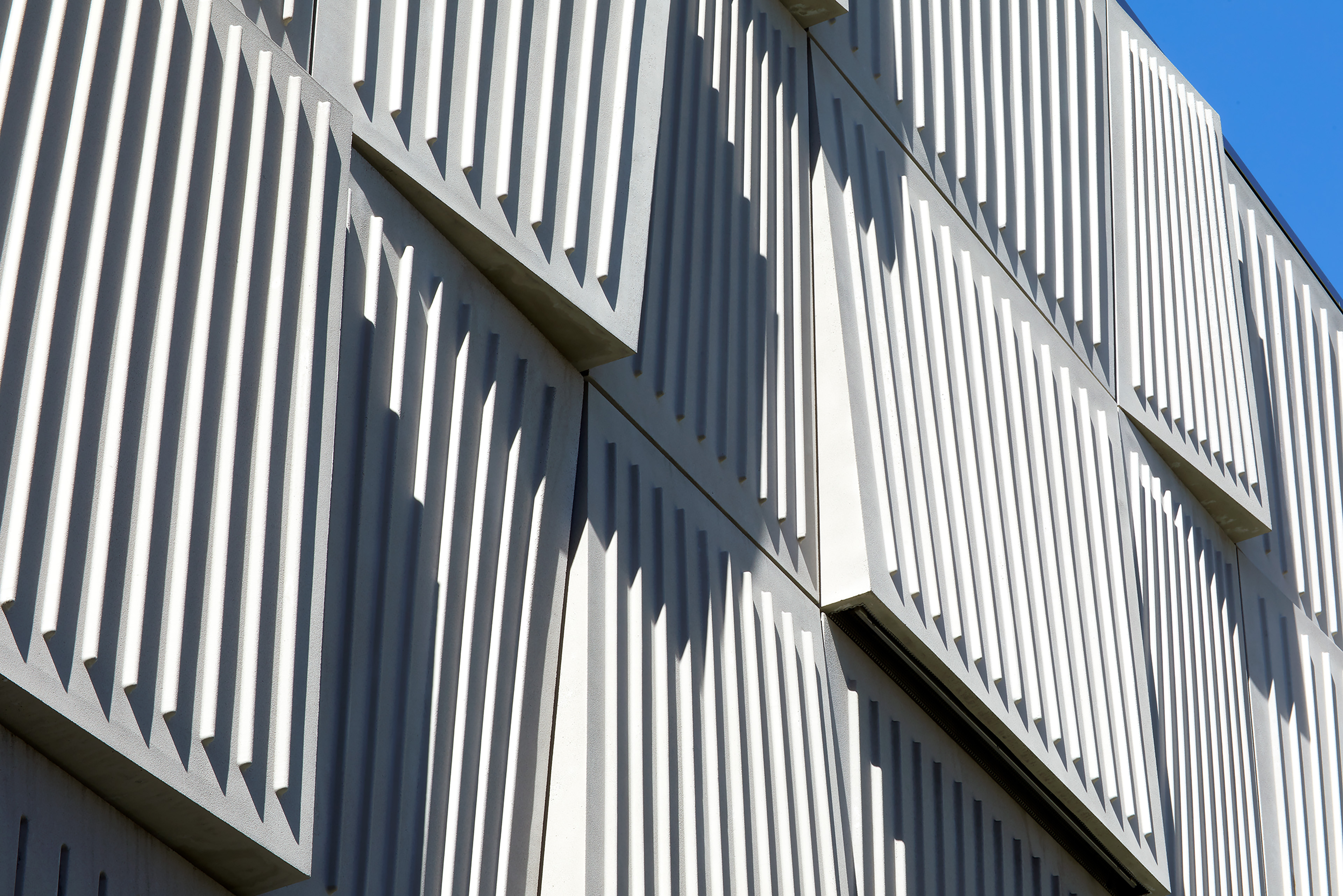 Close-up of angled, ribbed metal façade panels casting deep geometric shadows in bright sunlight.