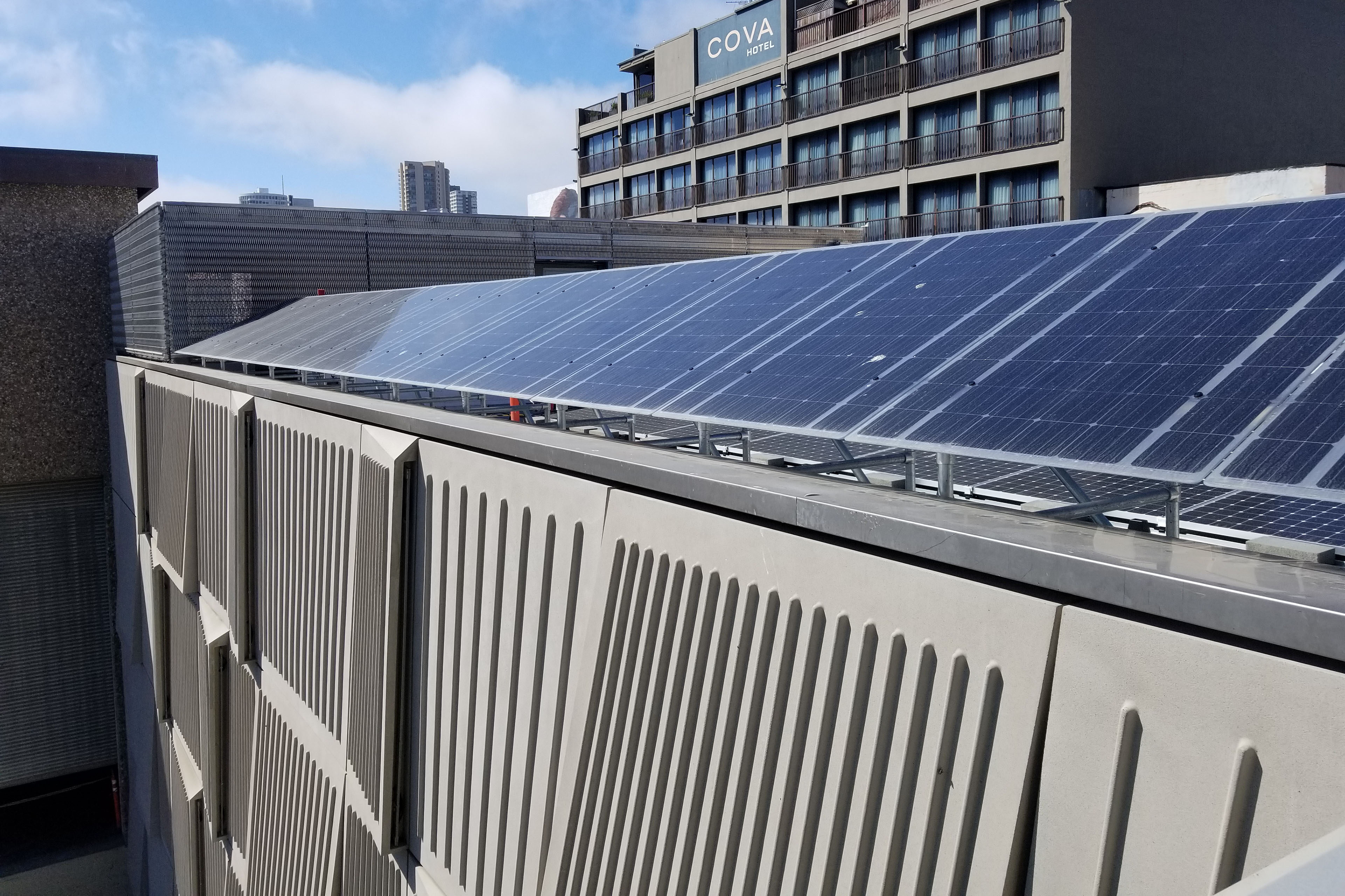 Solar panels mounted above a patterned metal building façade, with neighboring urban buildings visible in the background.
