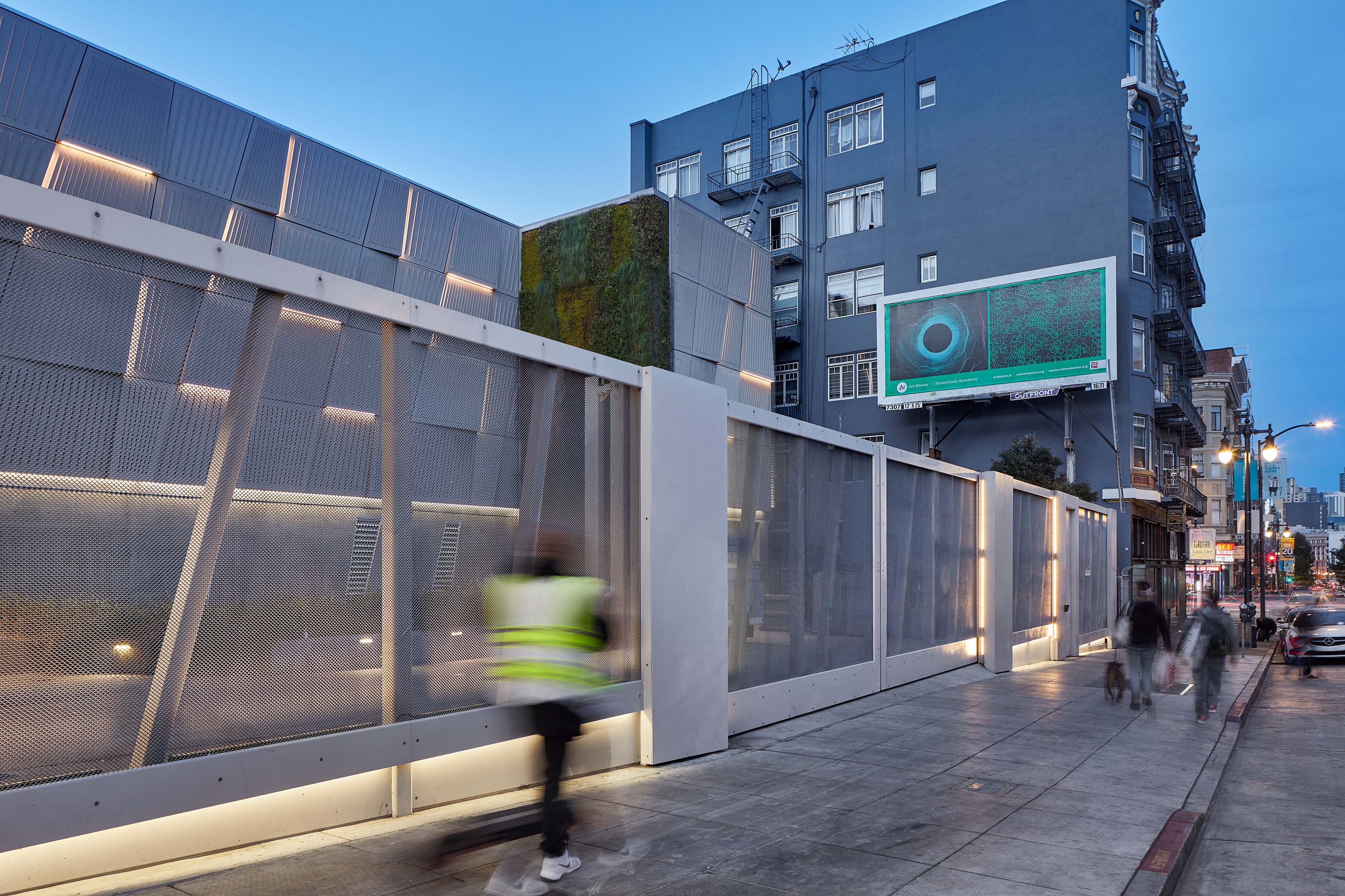 Street view of a utility building with perforated metal screening, integrated lighting, a vertical living wall, and pedestrians moving along the sidewalk.