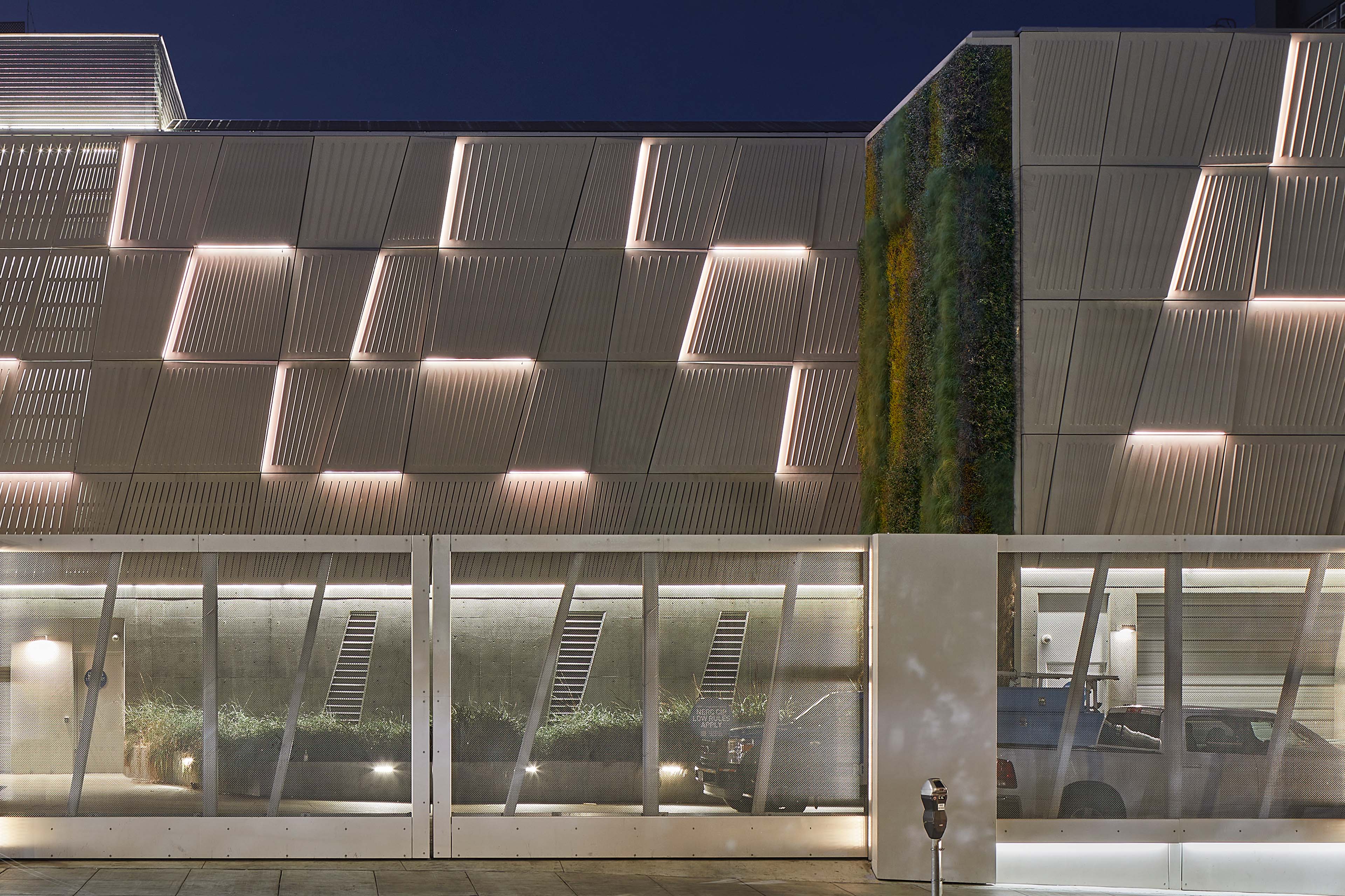 Illuminated utility building façade at dusk with patterned metal panels, a vertical living wall, and a screened service area along the sidewalk.