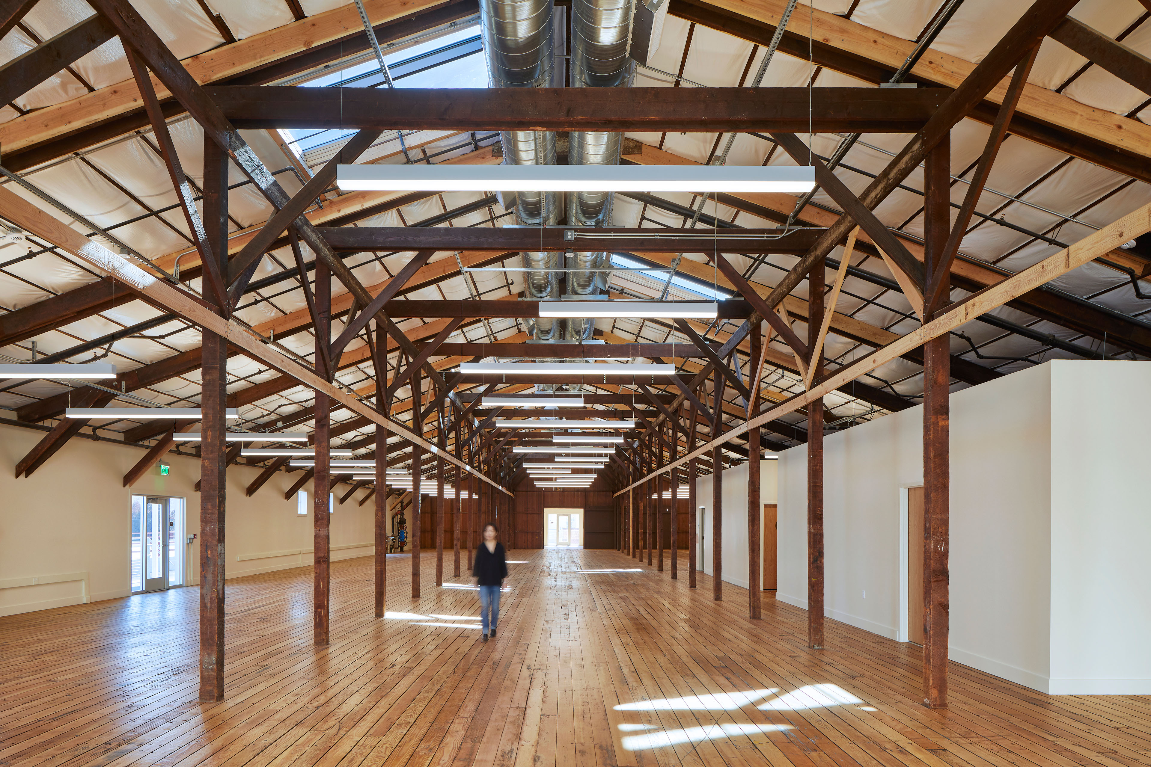 Long open interior with restored wood floors, exposed timber trusses, skylights, suspended linear lights, and visible ductwork, with one person walking through the space.