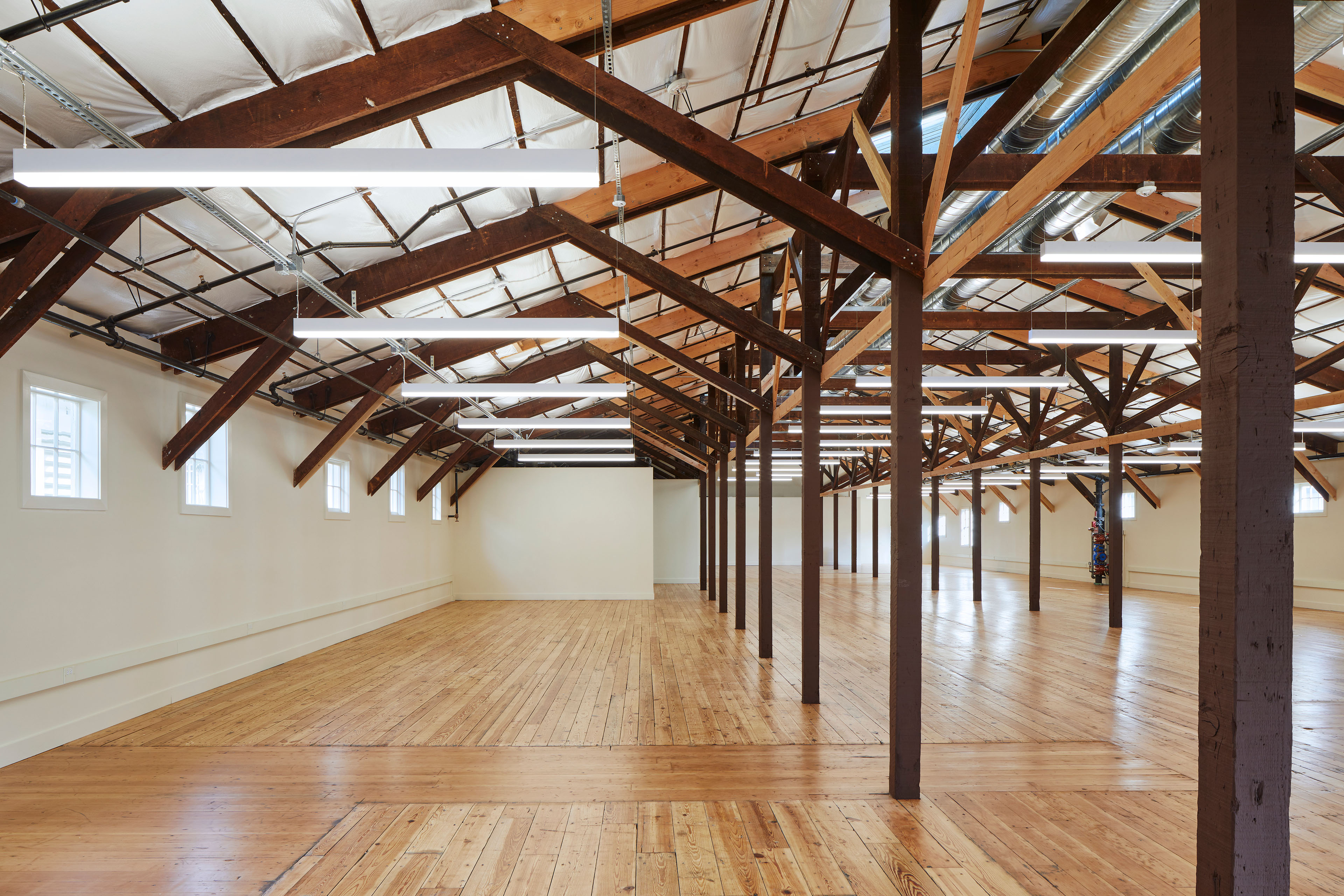 Expansive restored interior with wood floors, exposed timber columns and roof trusses, suspended linear lights, and visible ductwork.