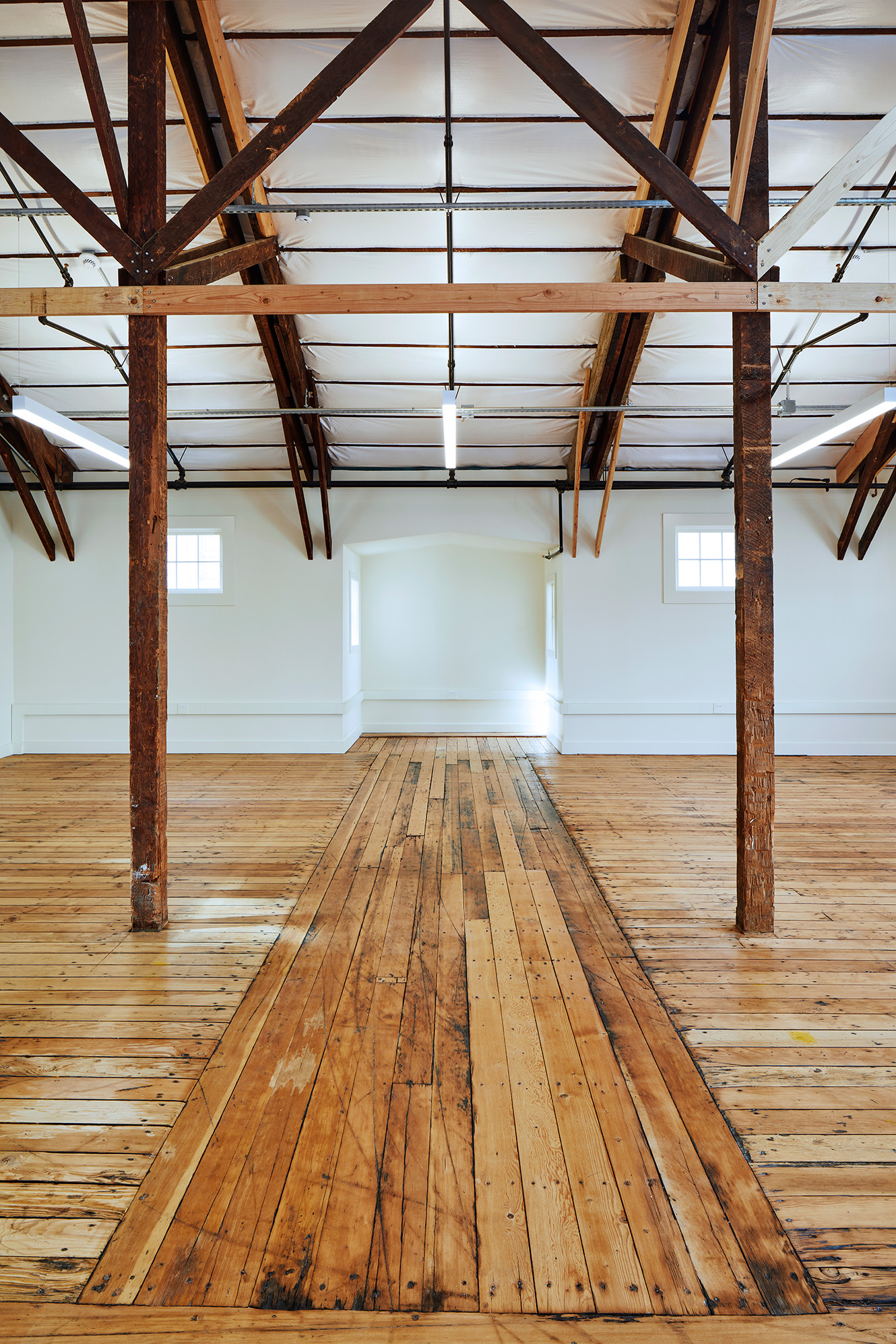 Restored open interior with wood floors, exposed timber columns and roof framing, white walls, and suspended linear lights.